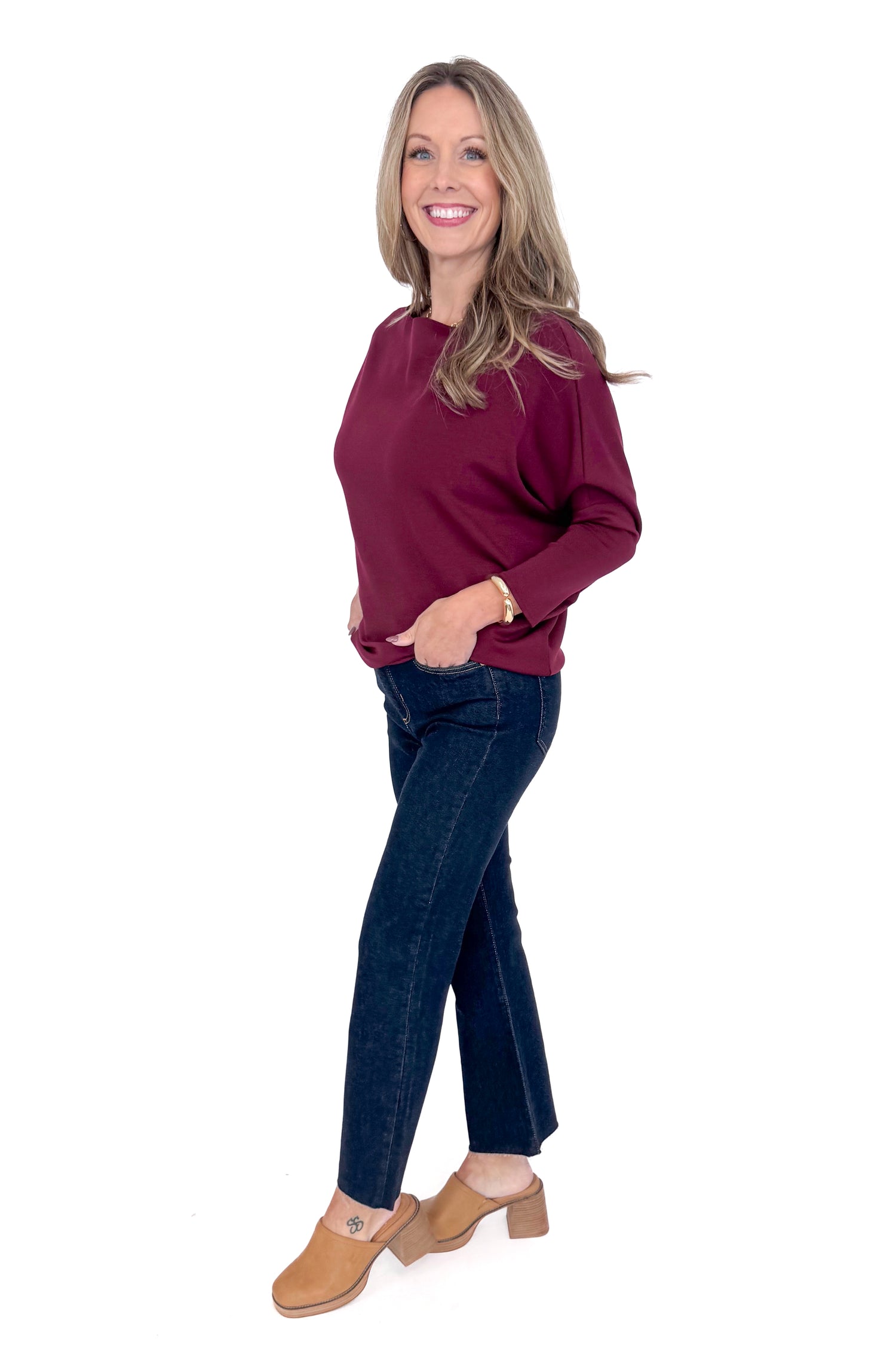 Side view of woman wearing burgundy top with jeans against white studio background.