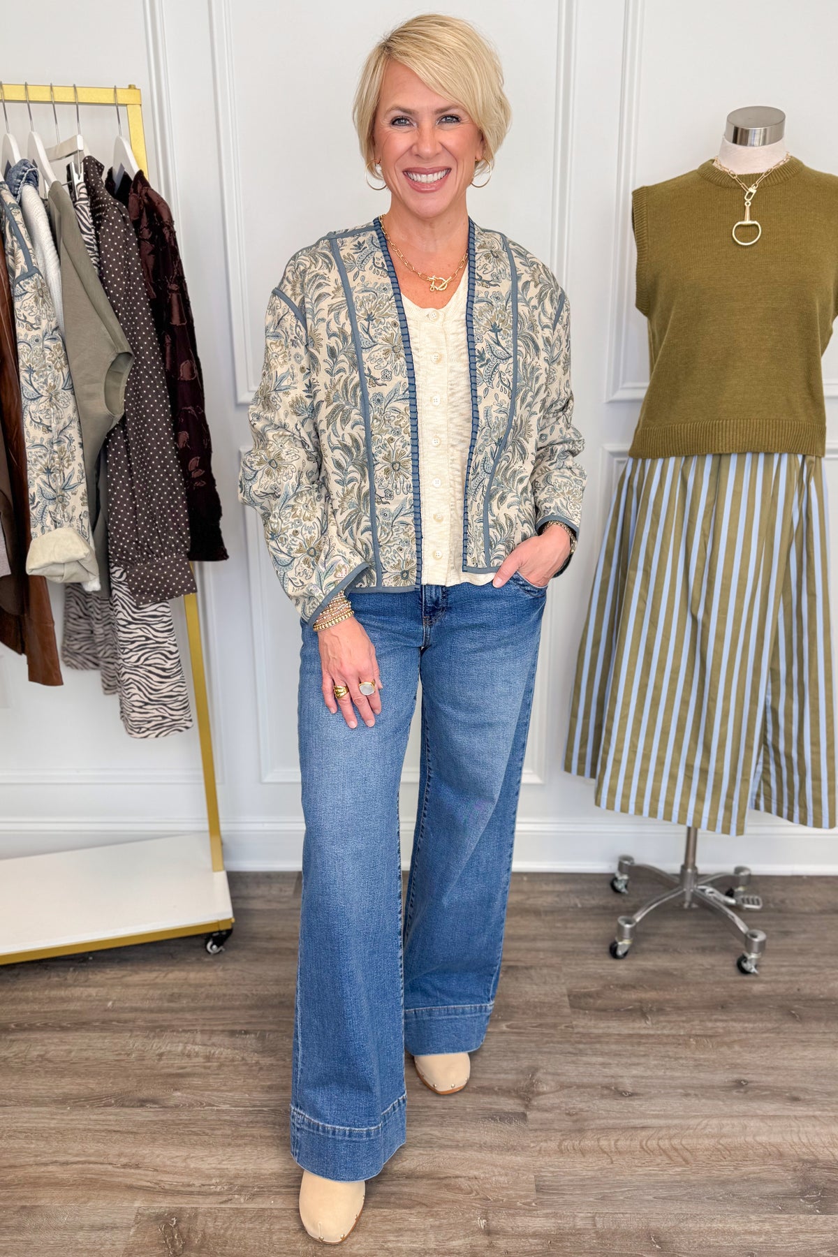 Front view of woman wearing Swagger Boutique’s Beige & Blue Printed Lightweight Jacket, Cream Sleeveless Button up Sweater with Maya Denim Jeans with clothing hanging in the background.