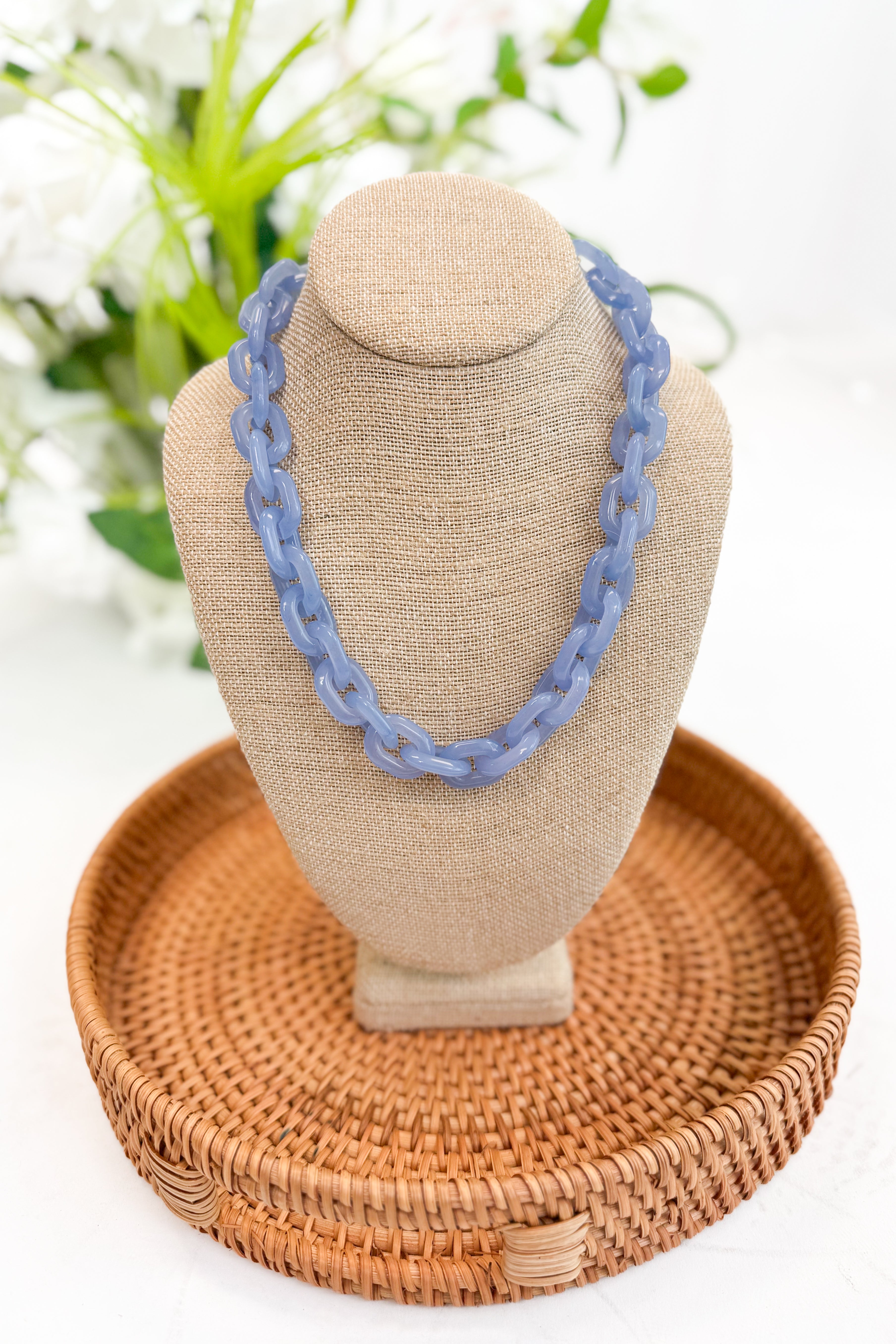 Blue necklace on a beige bust with a woven basket and blurred greenery in the background