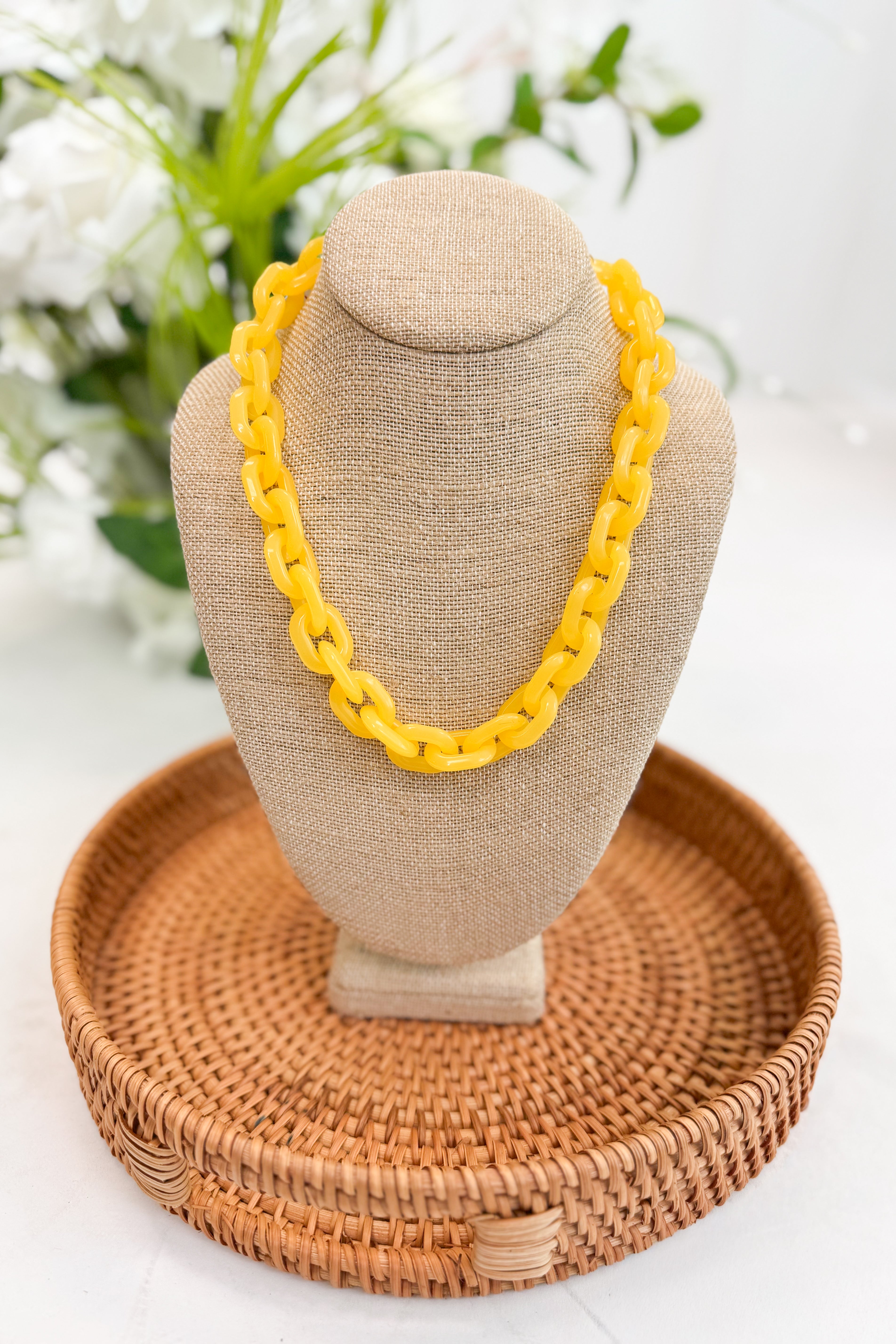 Yellow necklace on a beige mannequin head with a woven basket and blurred flowers in the background