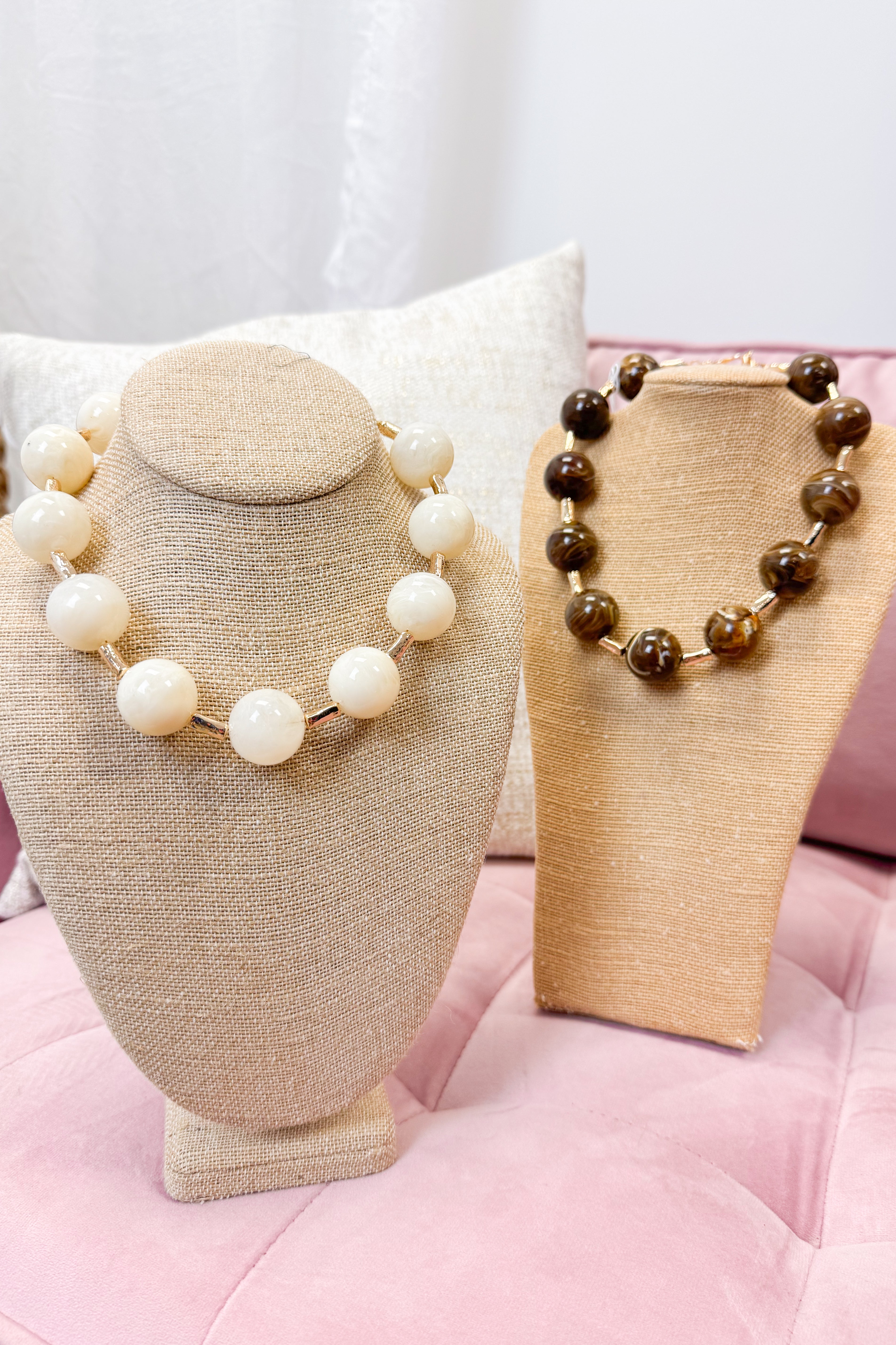 Two necklaces on mannequin heads with a pink cushion background