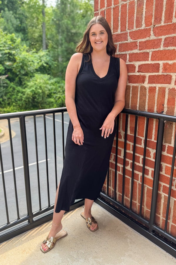Woman in a black dress standing on a balcony with a brick wall and greenery in the background