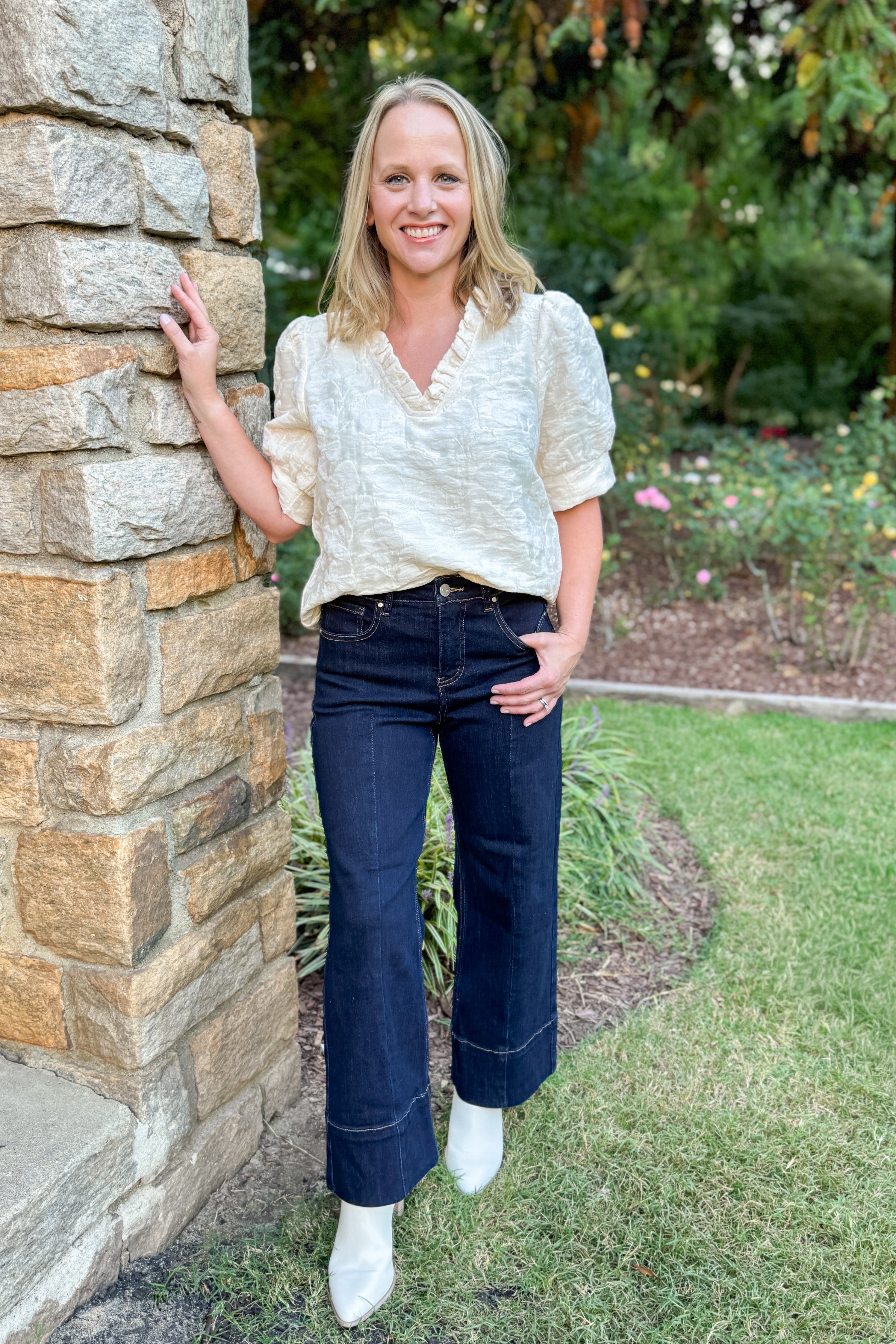 Front view of woman outside next to stone column wearing Cream Floral Textured V-neck Top and blue jeans.
