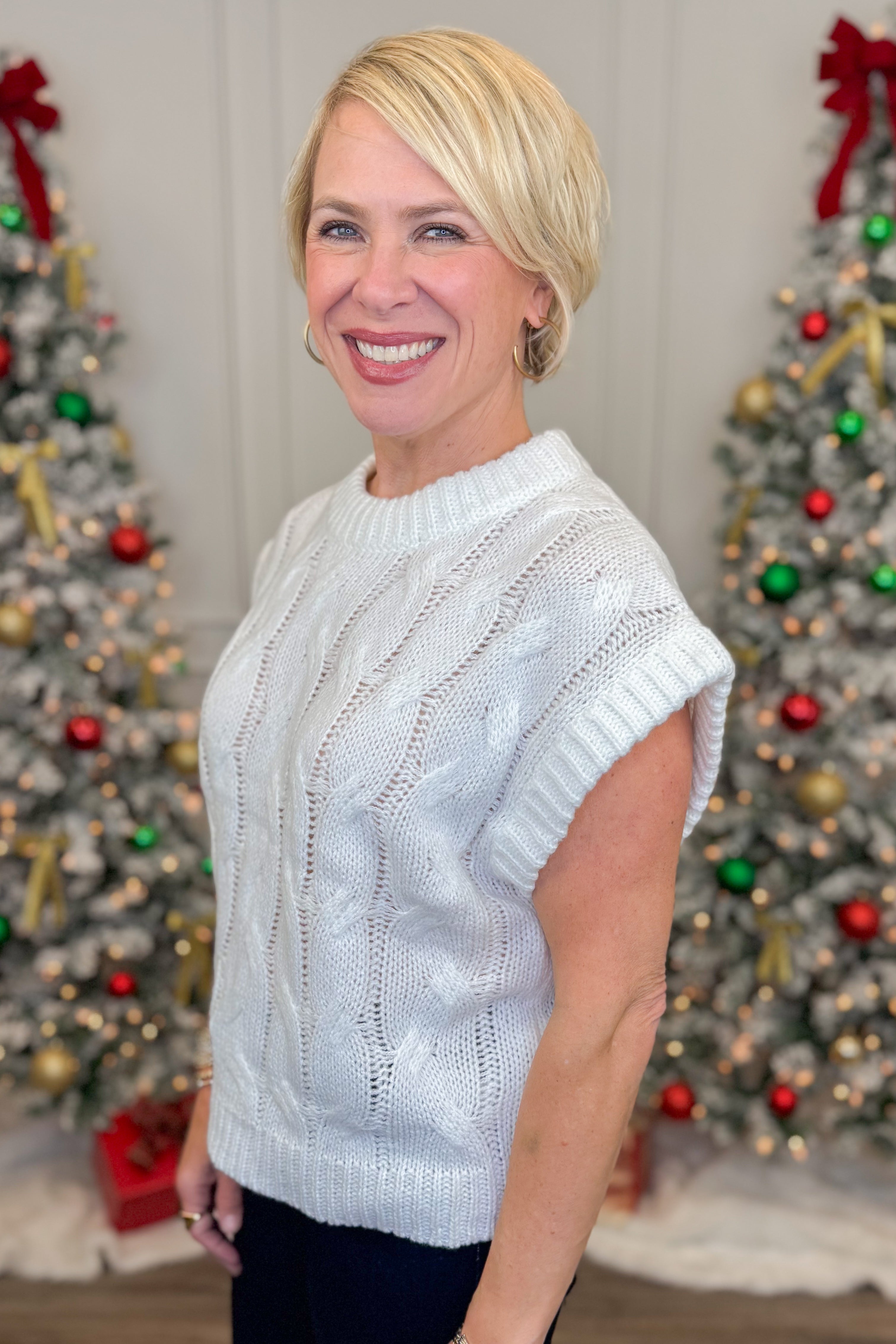 Woman wearing a white sweater standing in front of Christmas trees with decorations.