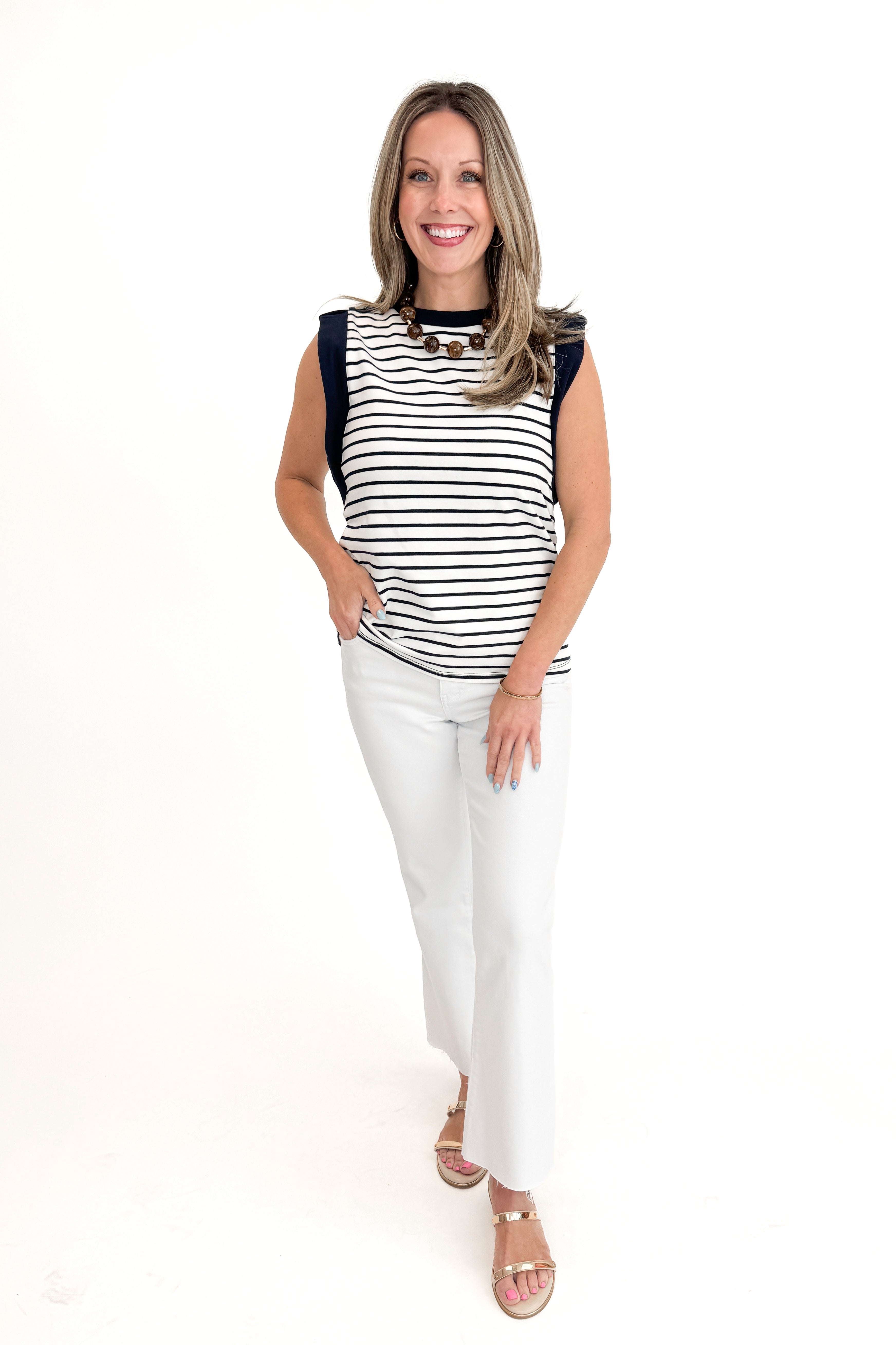 Front view of woman wearing White & Navy Striped Sleeveless Top with white jeans against white studio background.