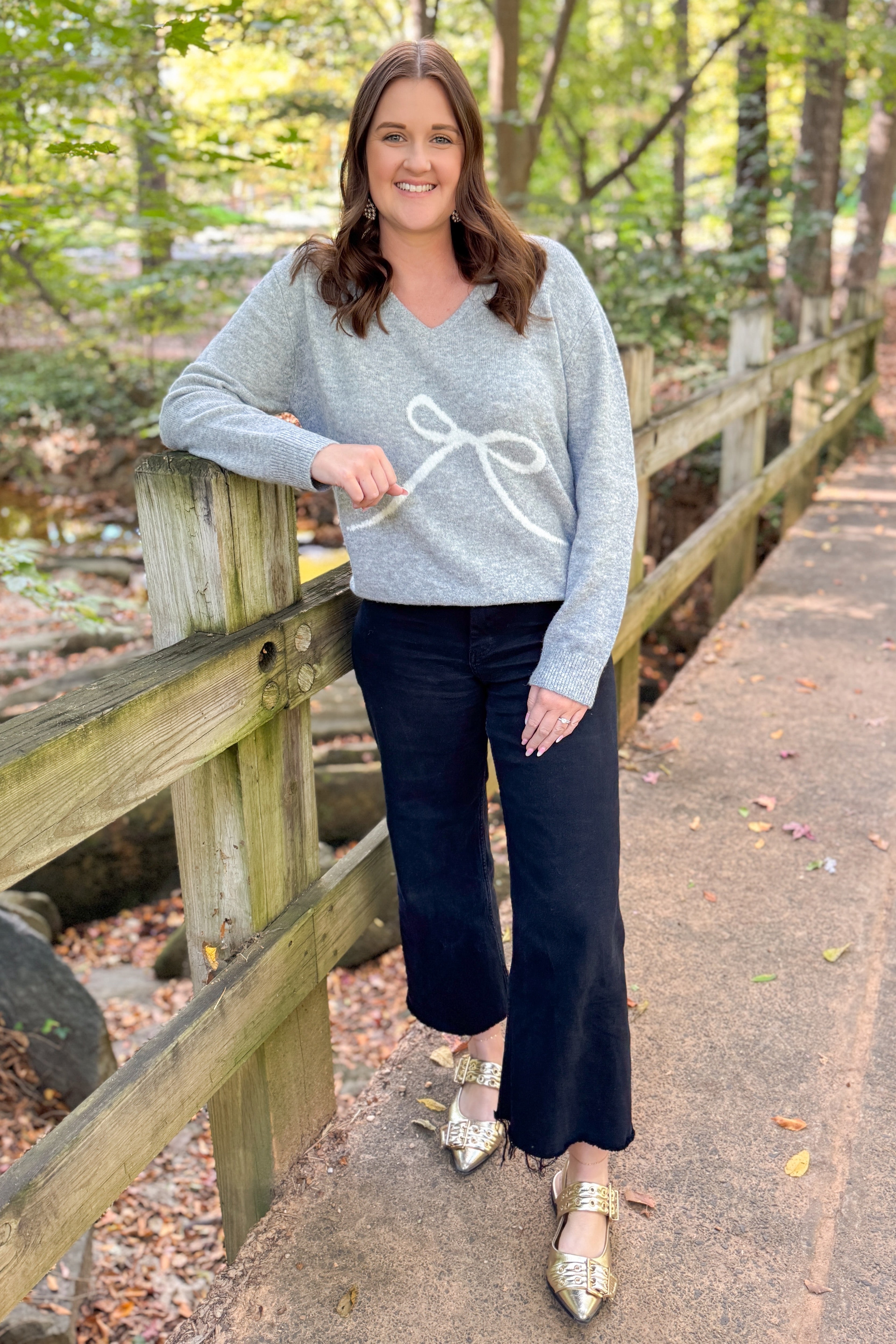 Front view of woman outdoor wearing Grey Bow Printed Sweater with jeans against with trees and leaves.
