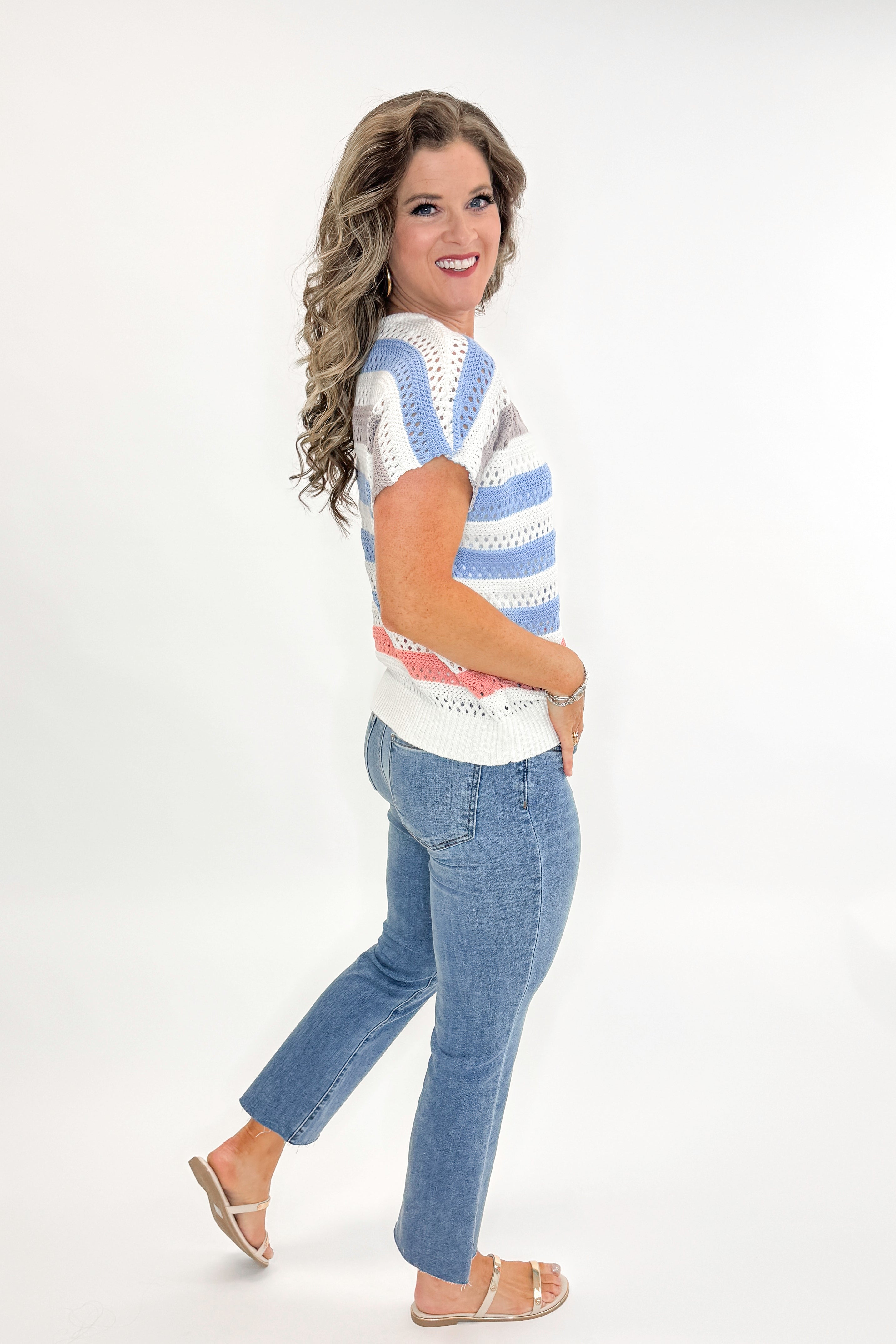 Side view of woman wearing Cream & Multi Striped Crochet Sleeveless Sweater with jeans against white studio background.