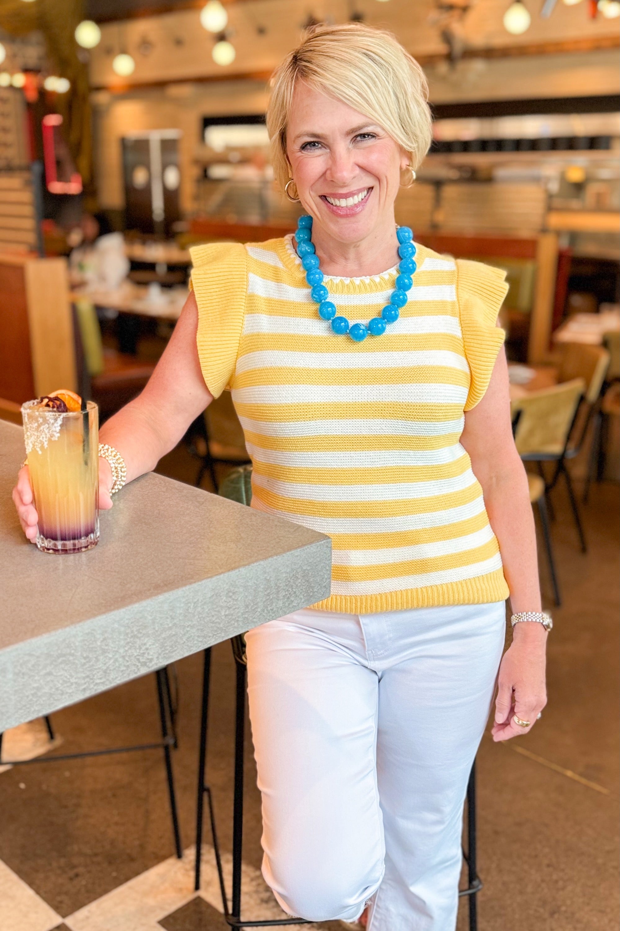 Front view of woman wearing Yellow Striped Flutter Short Sleeve Sweater with jeans standing at a restaurant bar.