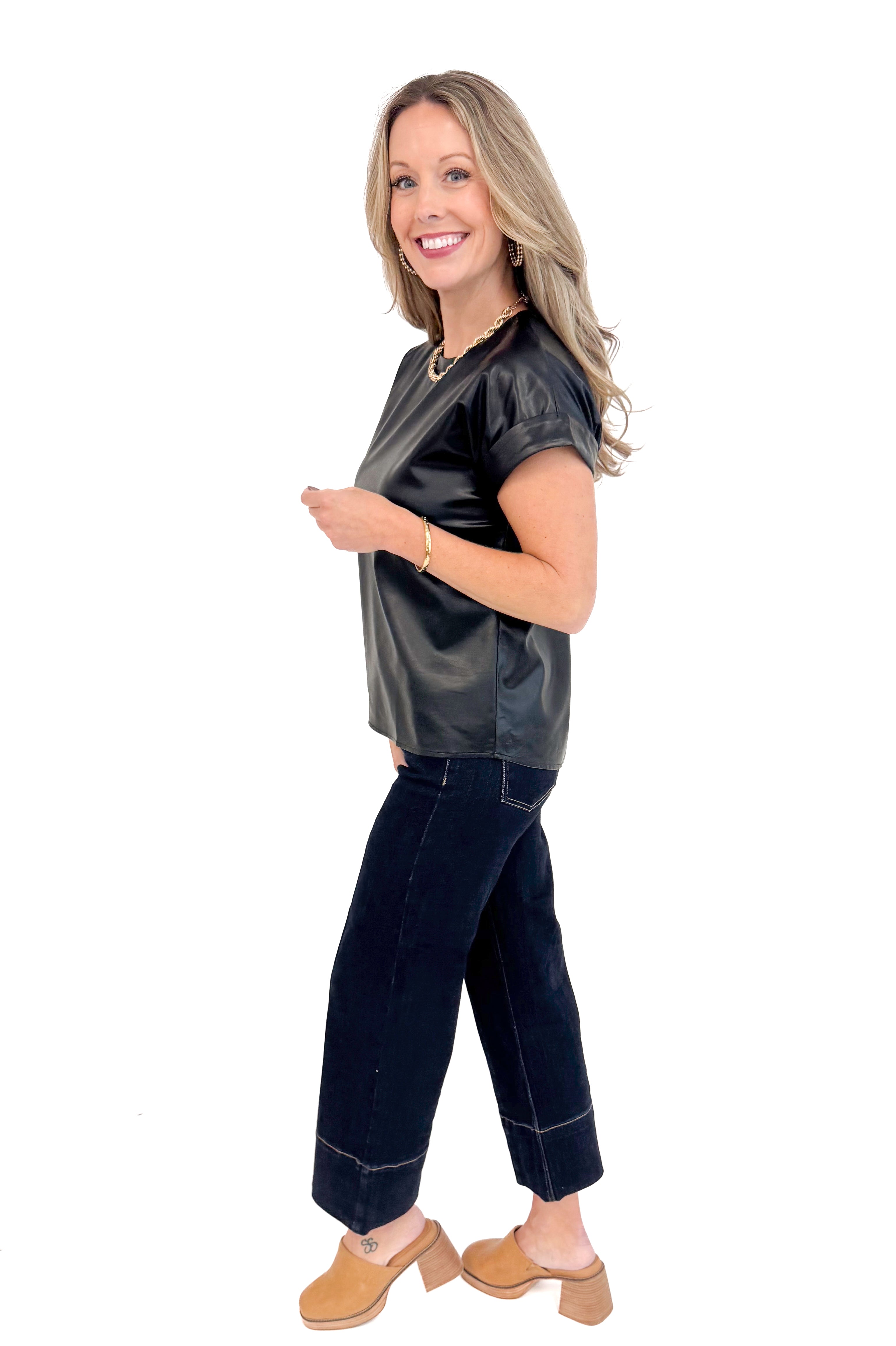 Full length side view of woman wearing black faux leather cap sleeve top with round neckline and blue jeans.