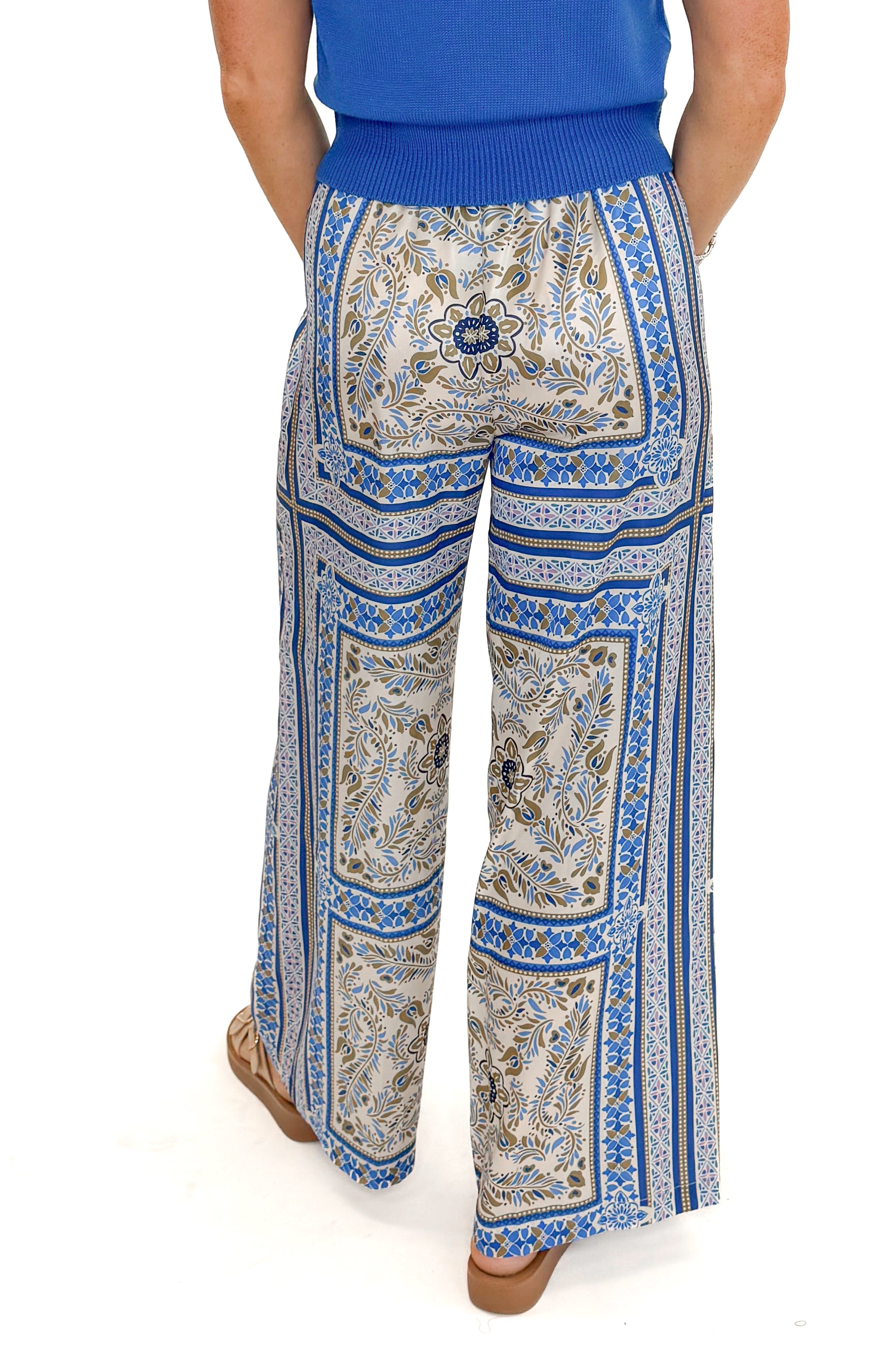 Back view of woman wearing Blue & Multi Scarf Print Pants by PINCH with blue sweater against white studio background.