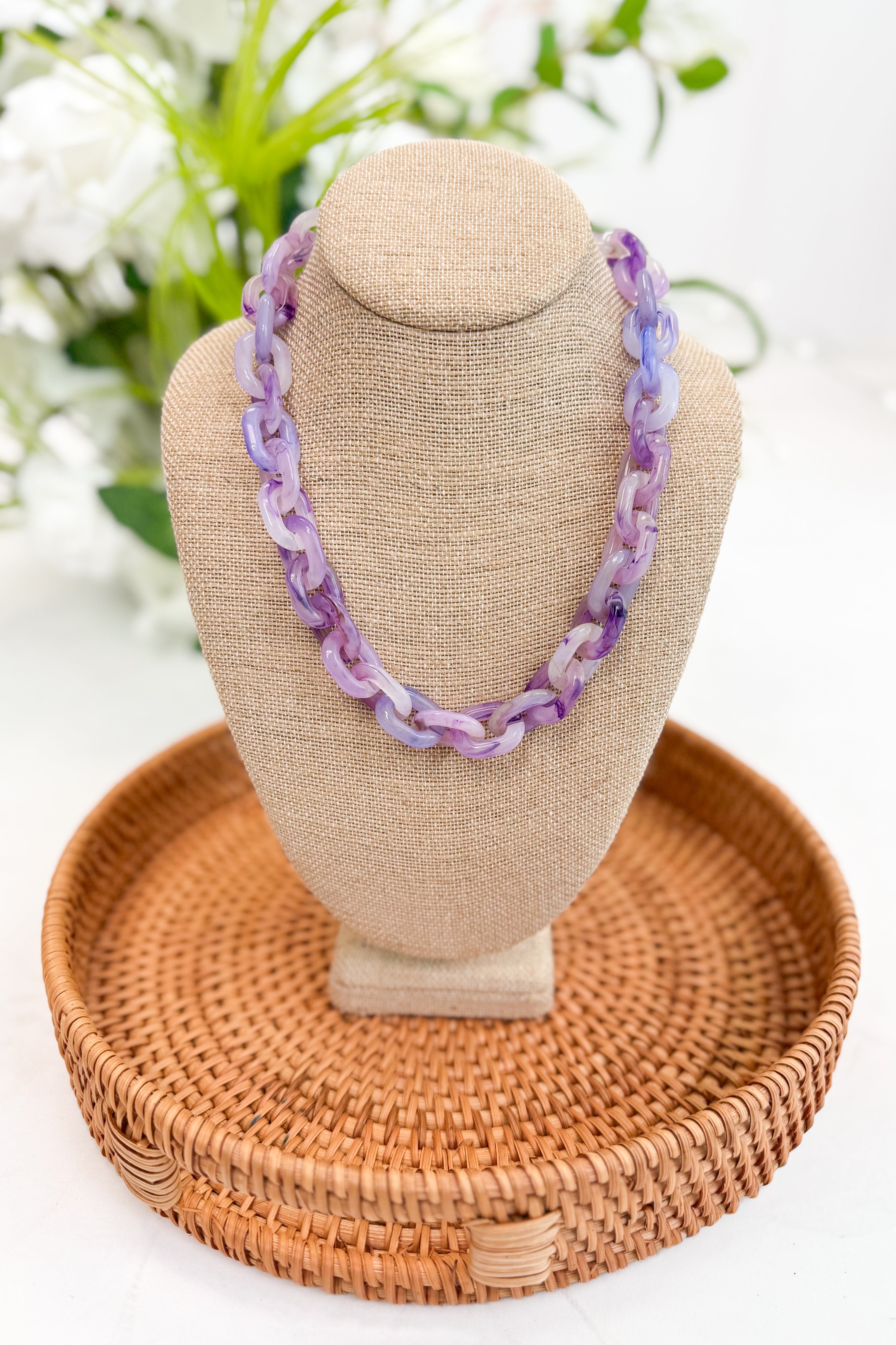 Necklace on a beige bust with a woven basket stand, against a blurred floral background