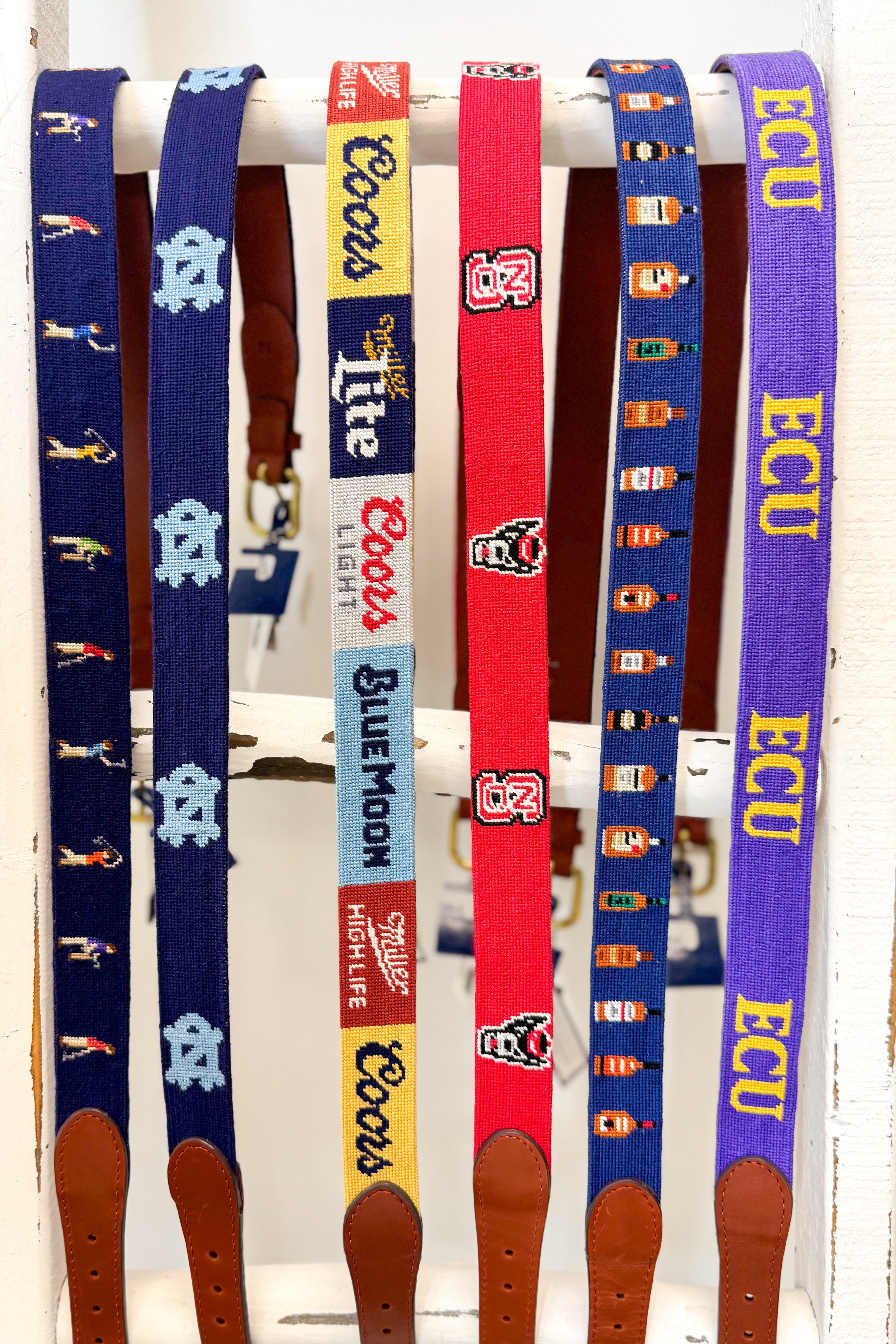 Six colorful embroidered utility belts with brown leather ends hanging vertically on a white rack. The belts feature various collegiate and brand logos, including Coors Light, Blue Moon, and ECU.