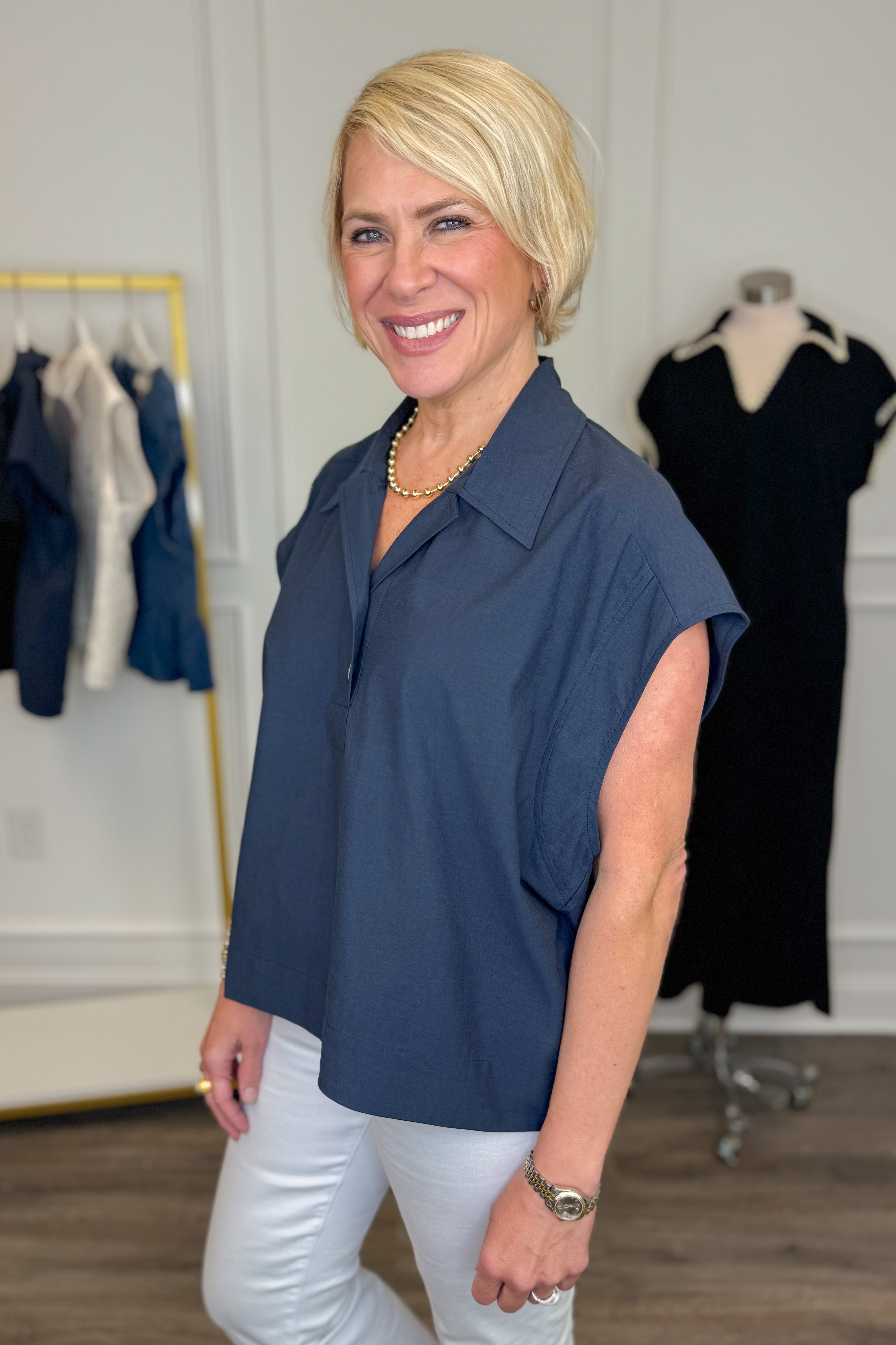 Side view of woman wearing Swagger Boutiqueβs Navy Collared Cap Sleeve Top with jeans with clothing hanging in the background.