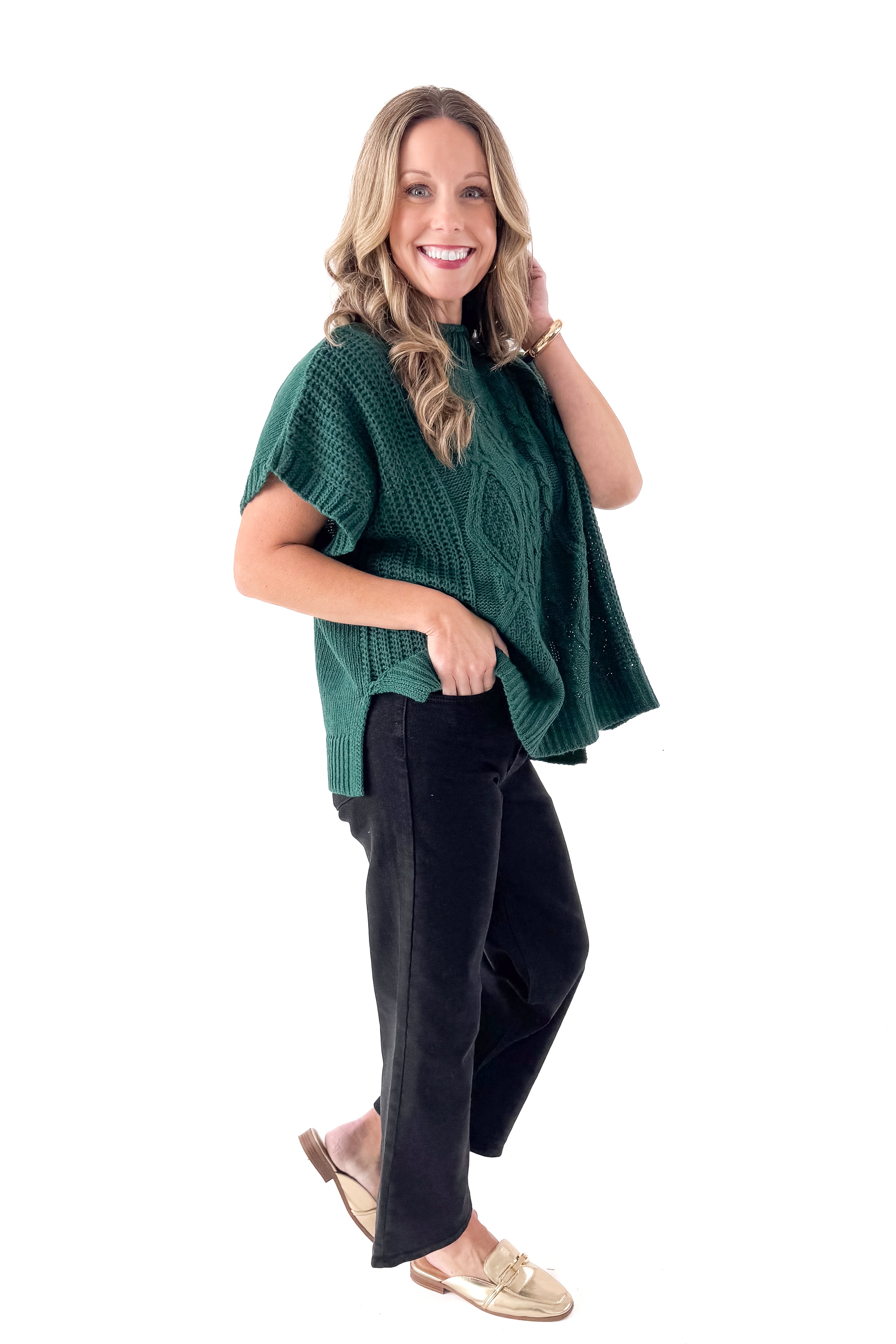 Side view of woman wearing Green Claude Cable Knit Sweater with jeans against white studio background.