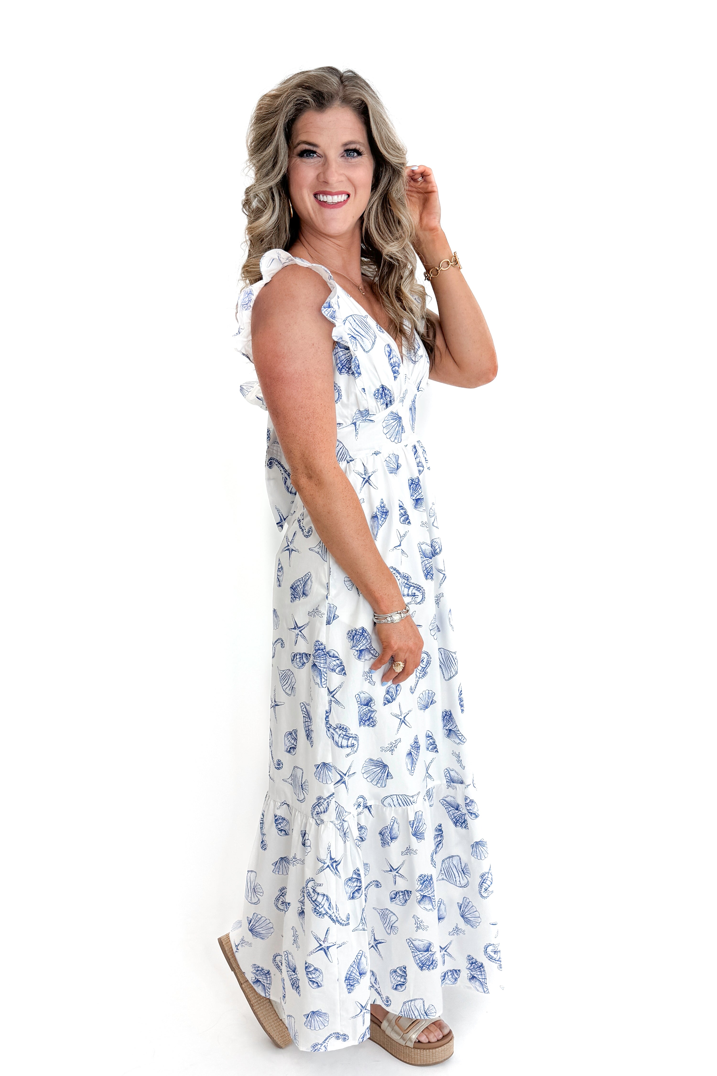 Side view of woman wearing White & Blue Shell Print Tie Back Maxi Dress by Skies Are Blue against white studio background.