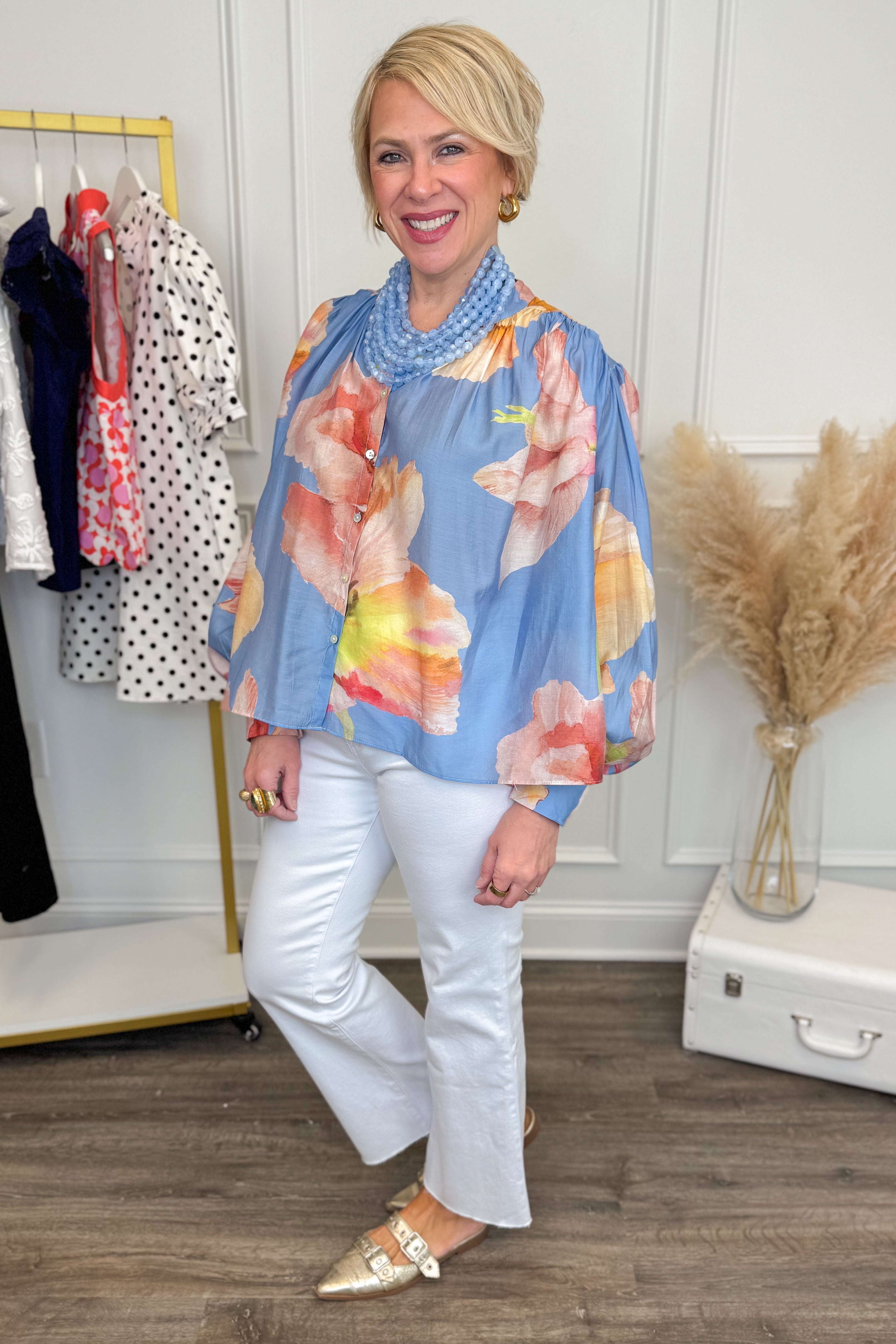 Woman wearing a floral blouse and white pants in a room with clothing racks.
