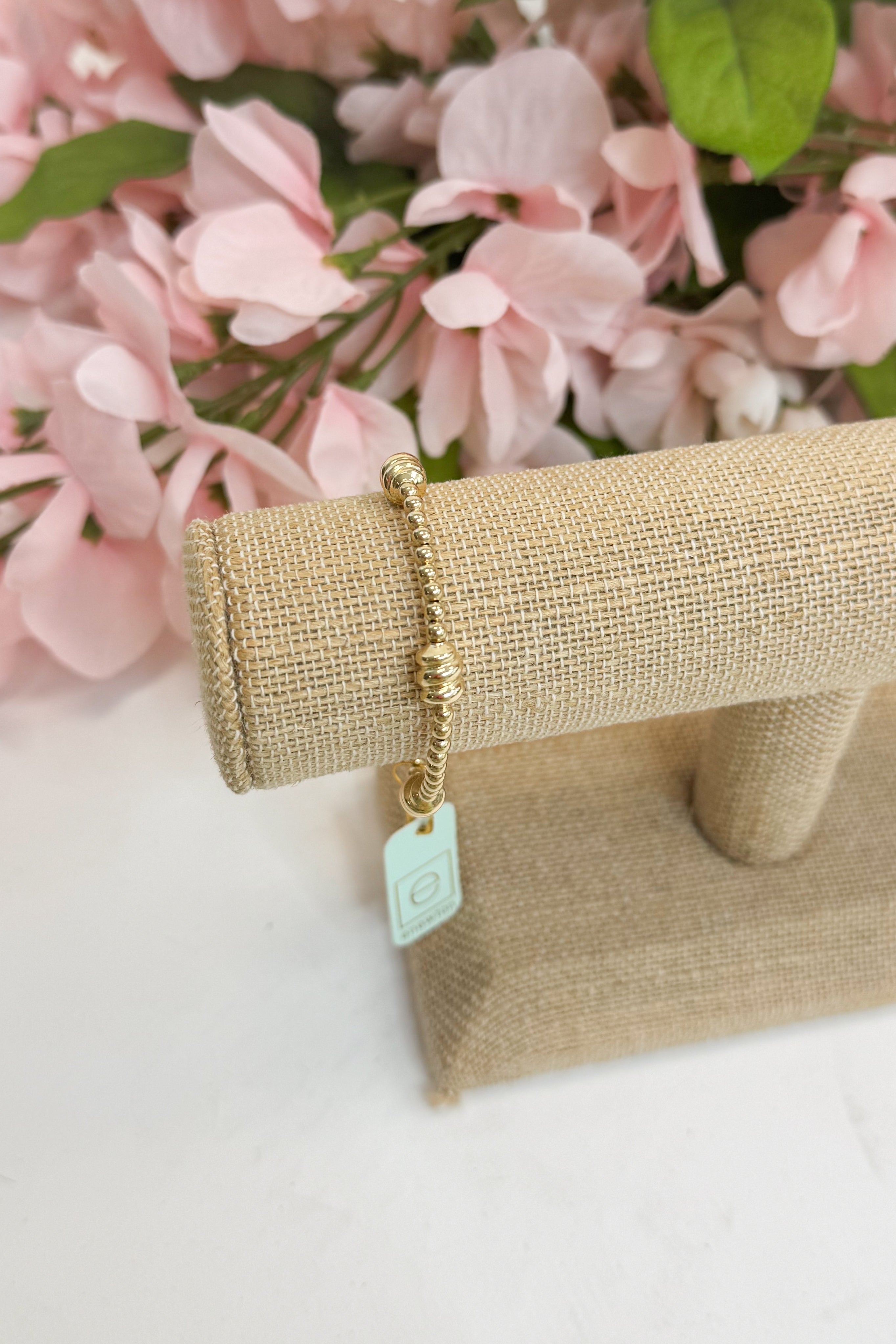 Gold bracelet with a tag on a beige stand against a pink floral background