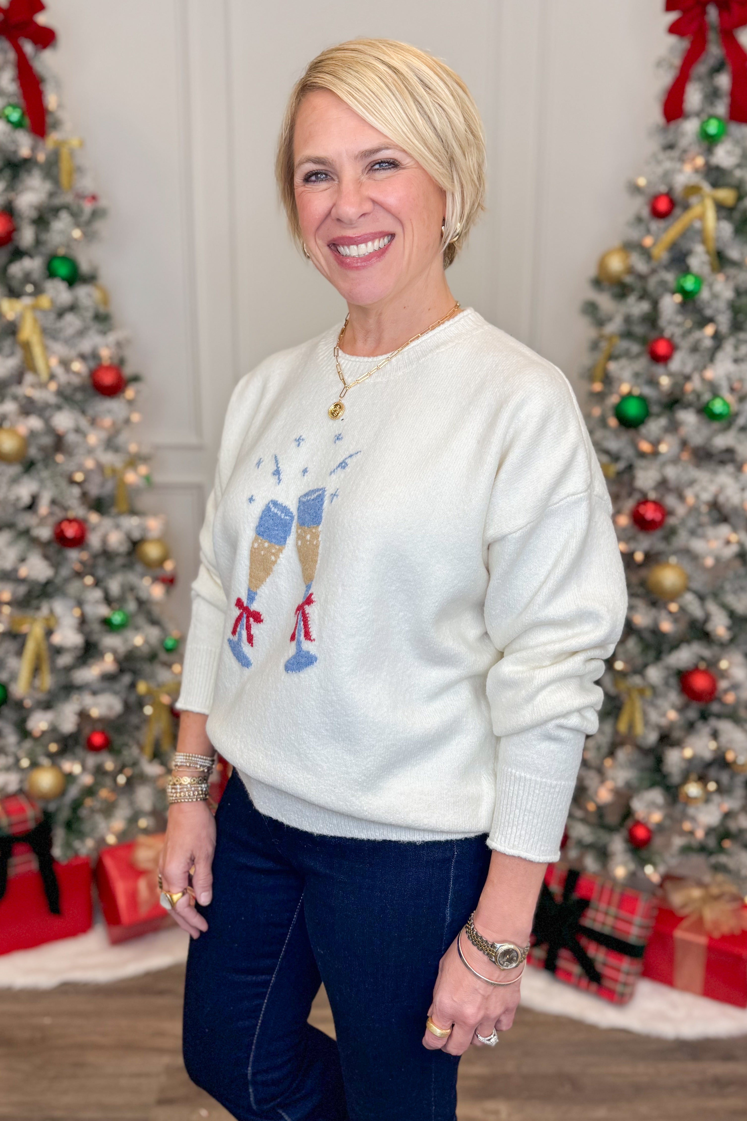 Woman wearing a white sweater with a graphic design in front of Christmas trees.