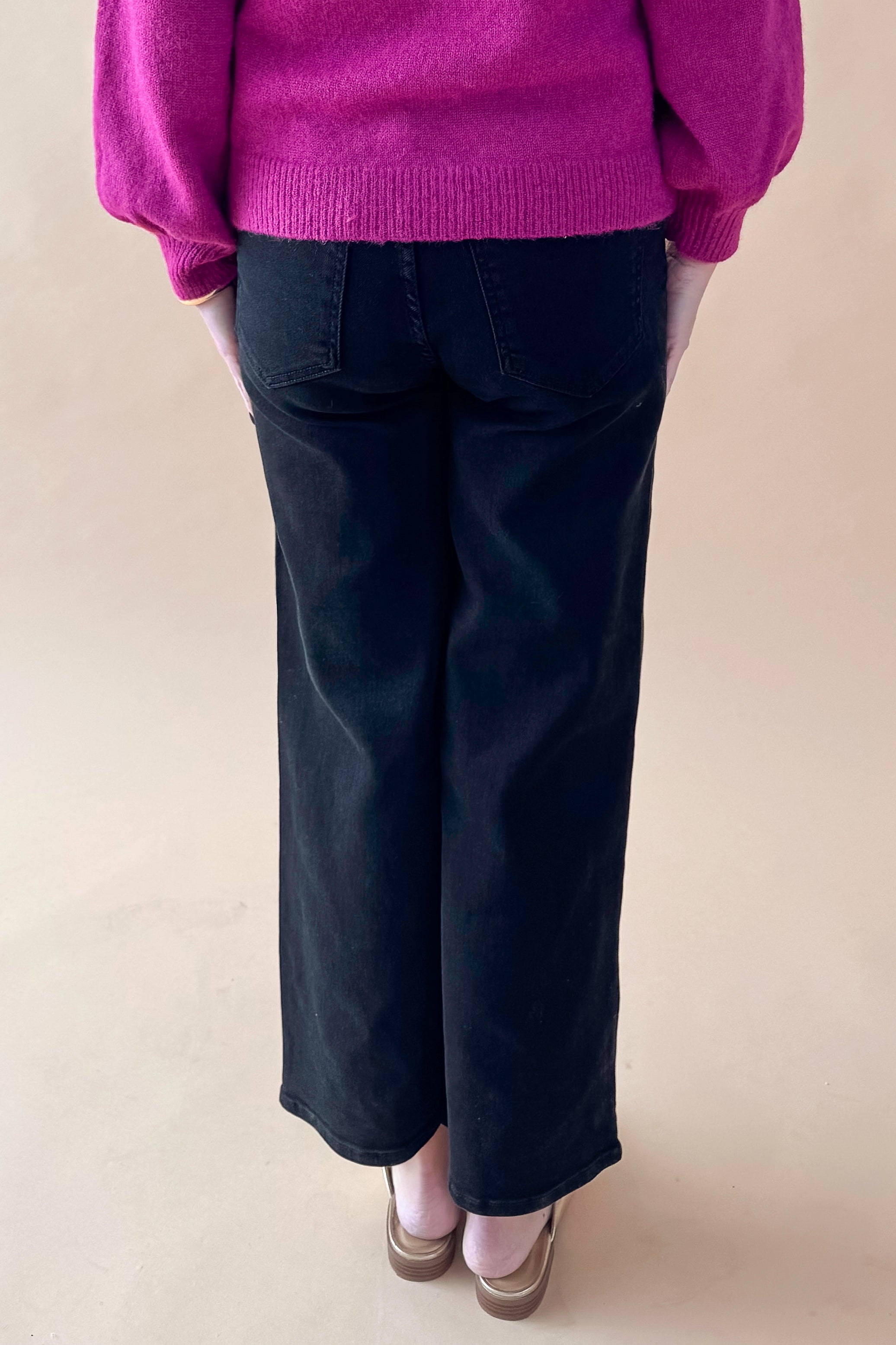 Back view of woman wearing purple top with black wide leg jeans against tan studio background.