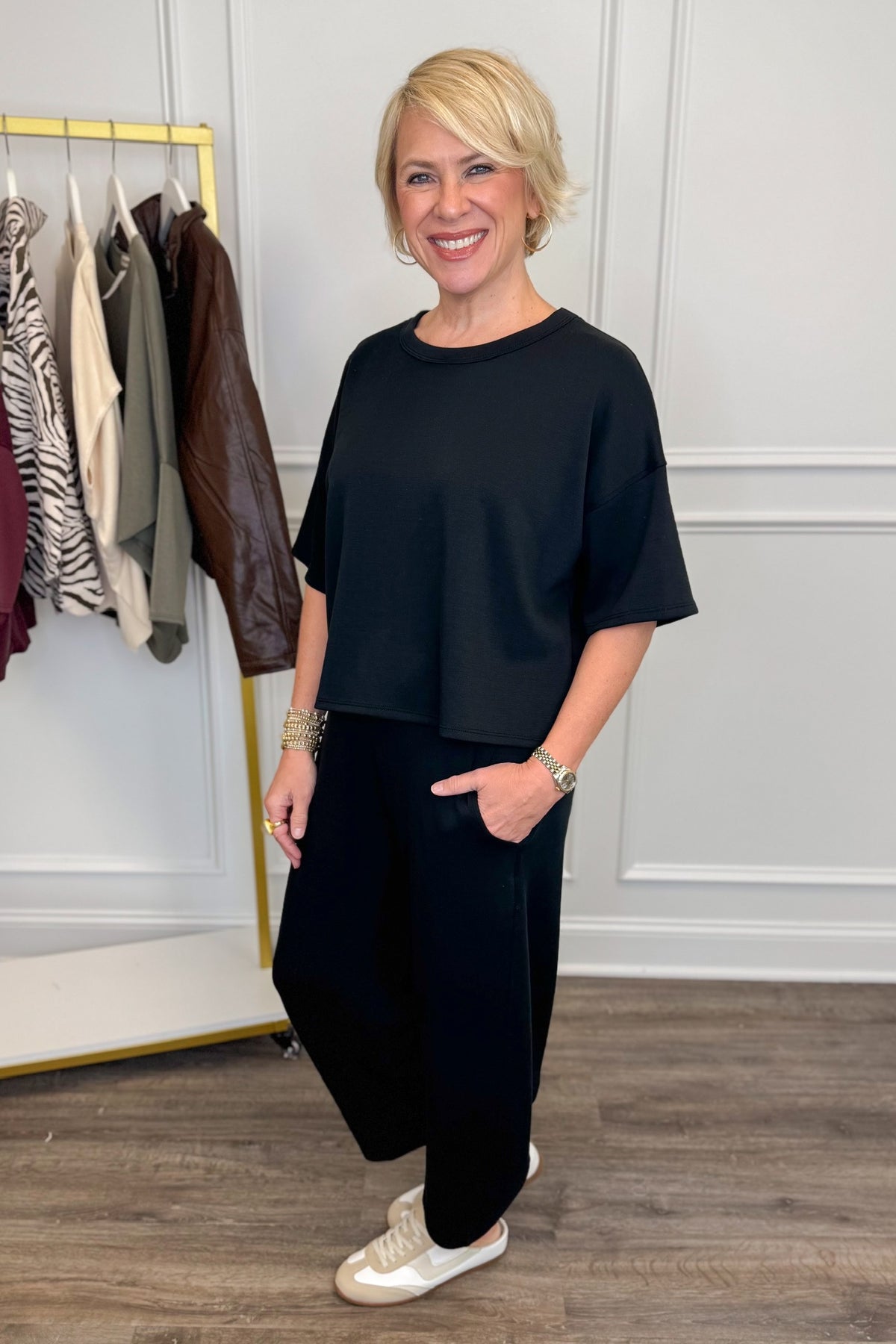 Side view of woman wearing Swagger Boutique’s Black Round Neck Short Sleeve Top with matching barrel pants with clothes hanging in the background.