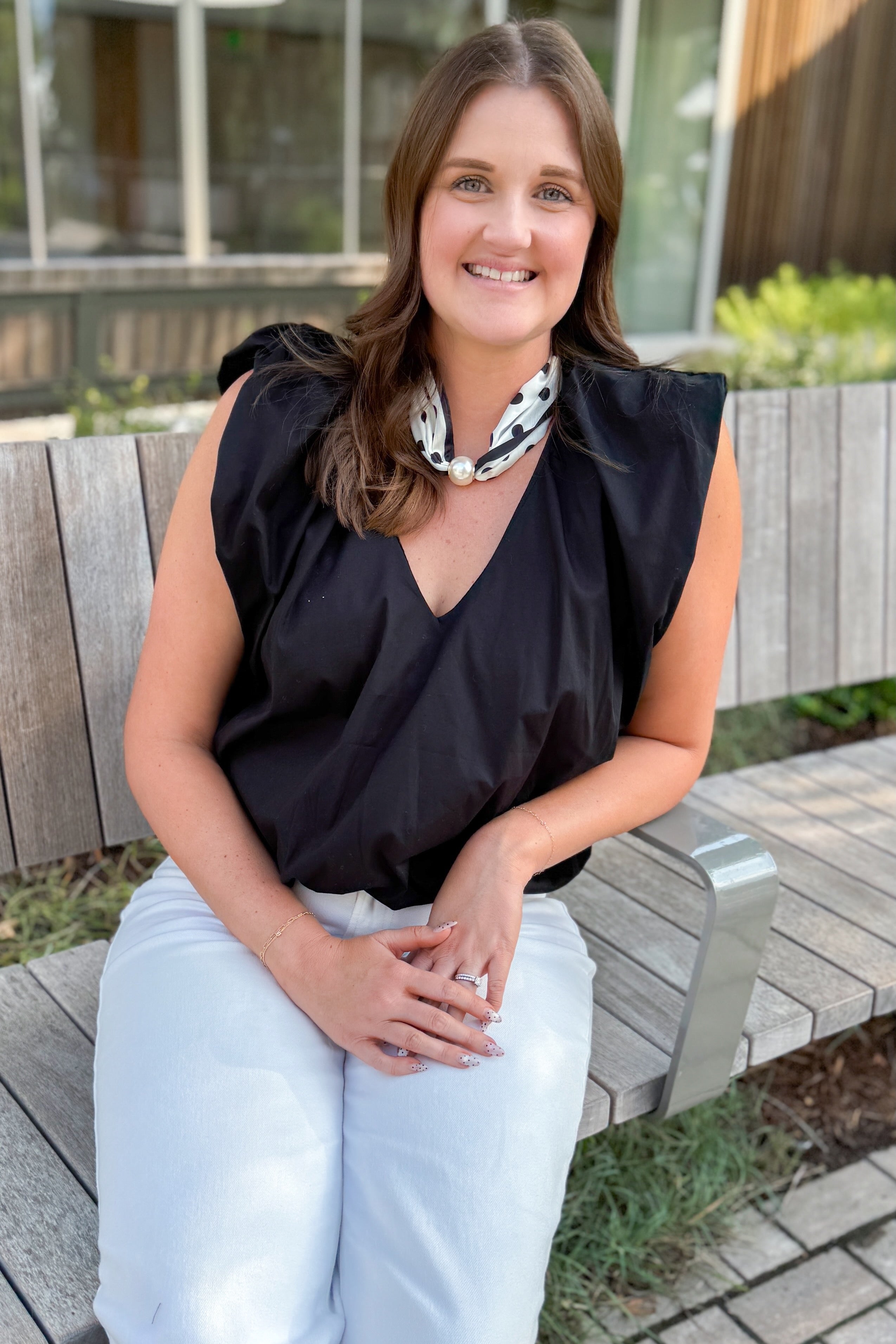 Front view of woman wearing Swagger Boutique’s Black Bubble Hem Pleated Top with jeans sitting outside on a bench.