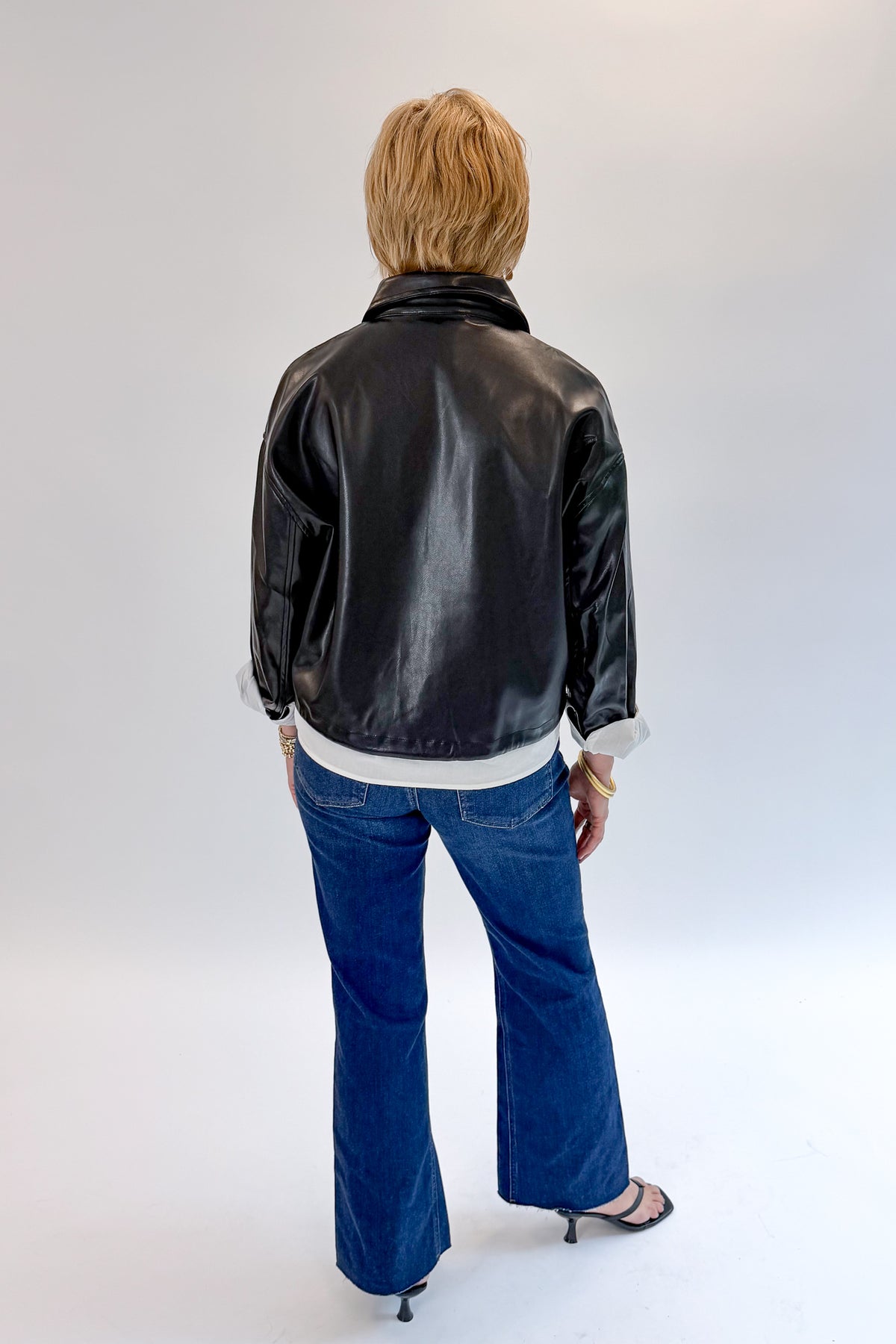 Back view of woman wearing Swagger Boutique’s Black Faux Leather Jacket With Layered Shirt with jeans against white studio background.