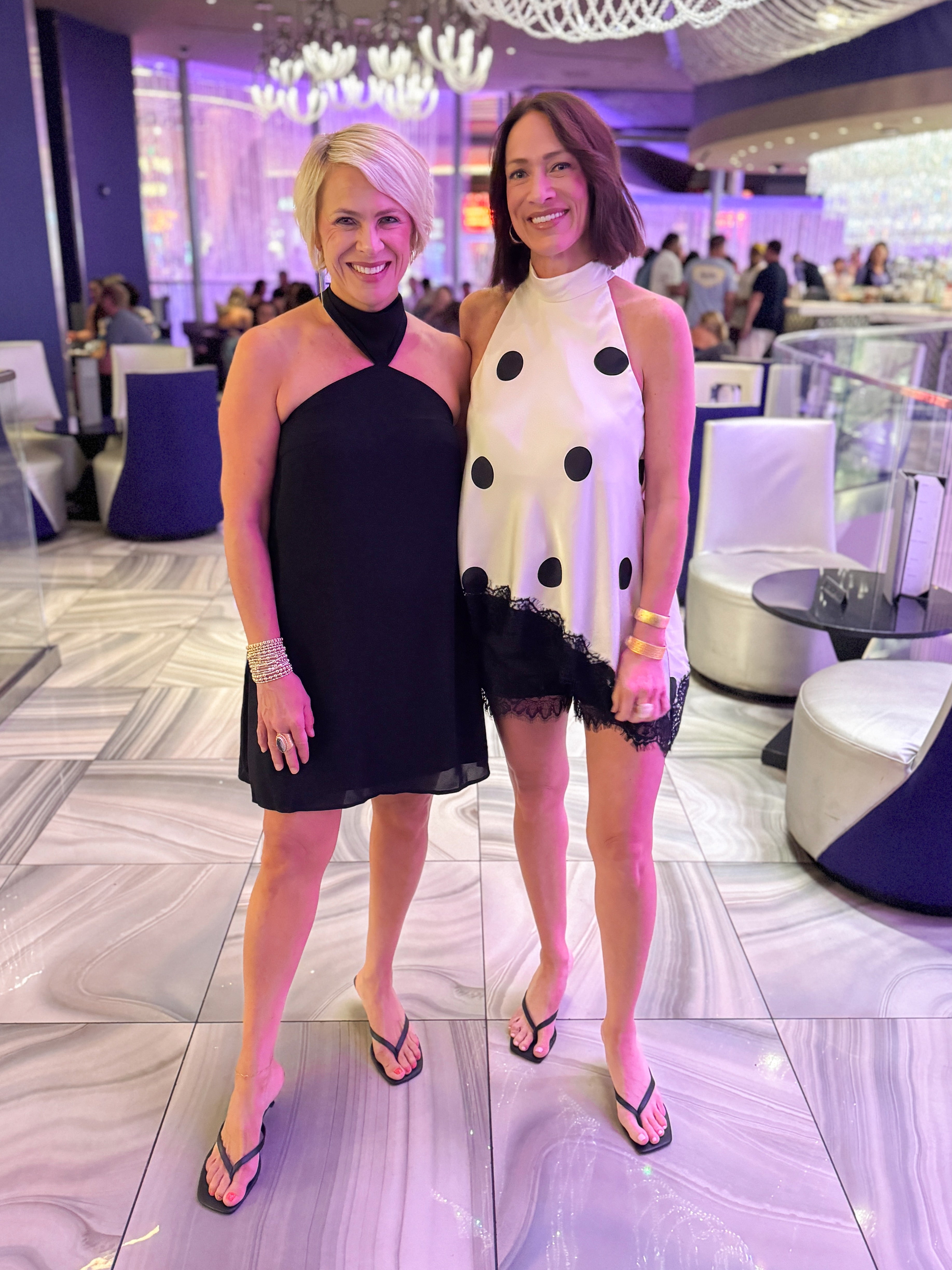 Front view of women wearing Swagger Boutiqueās Black Halter Neck Mini Dress with heels and polka dot halter top at a fancy restaurant.