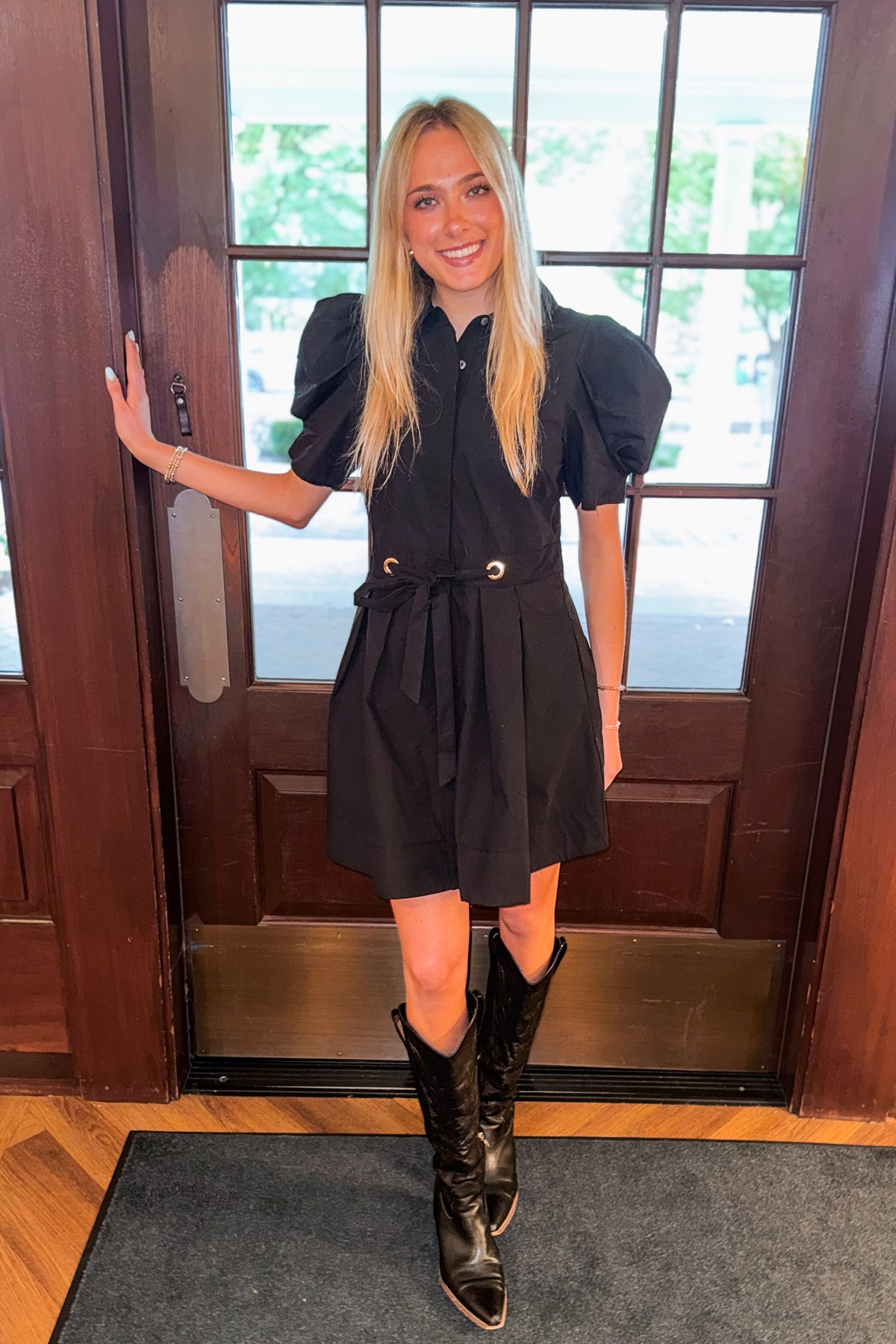 Front view of woman wearing Swagger Boutique’s Black Puff Sleeve Mini Dress by Entro standing by a front door.