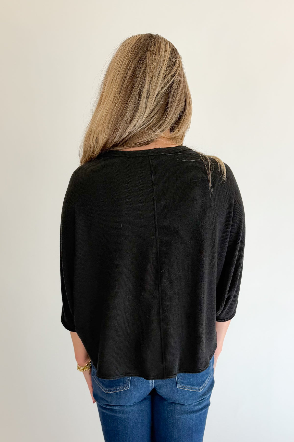 Back view of woman wearing Swagger Boutique’s Black Ribbed Dolman Sleeve Top with jeans against white studio background.