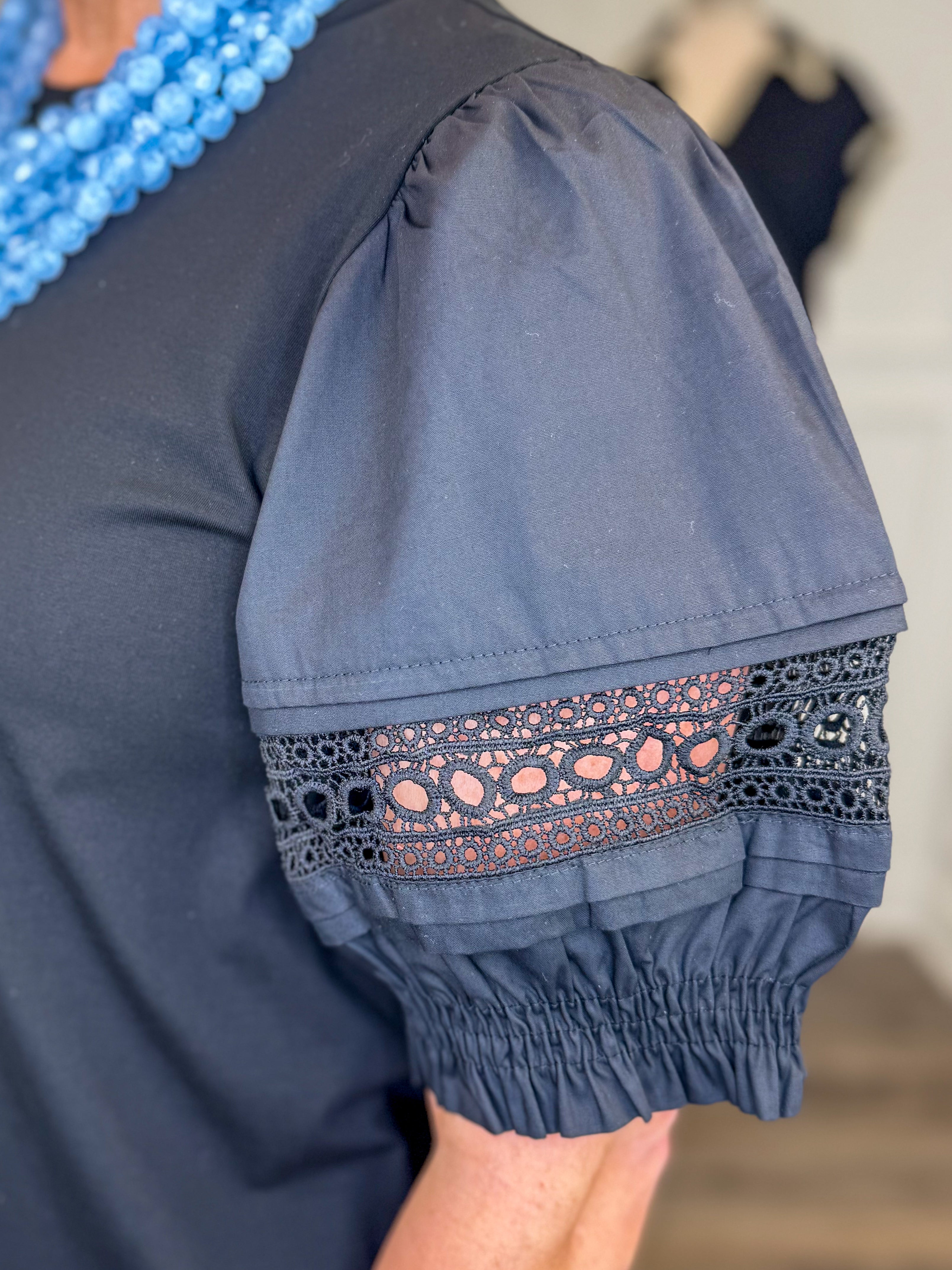 Front detail view of woman wearing Swagger Boutiqueβs Black Lace Sleeve Top with jeans with clothing hanging in the background.