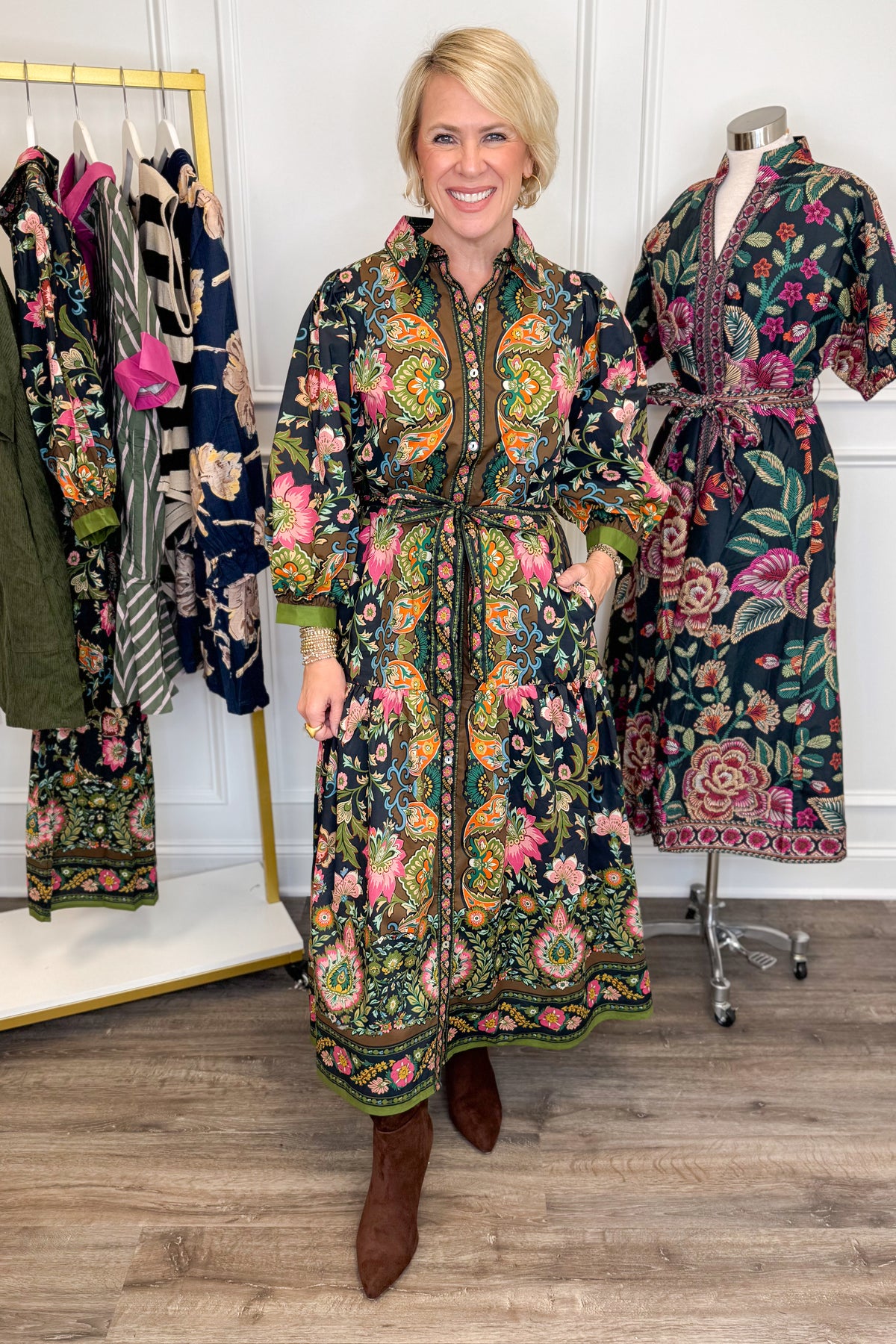 Front detail view of woman wearing Swagger Boutique’s black multi printed midi dress by Entro with boots with clothing hanging in the background.