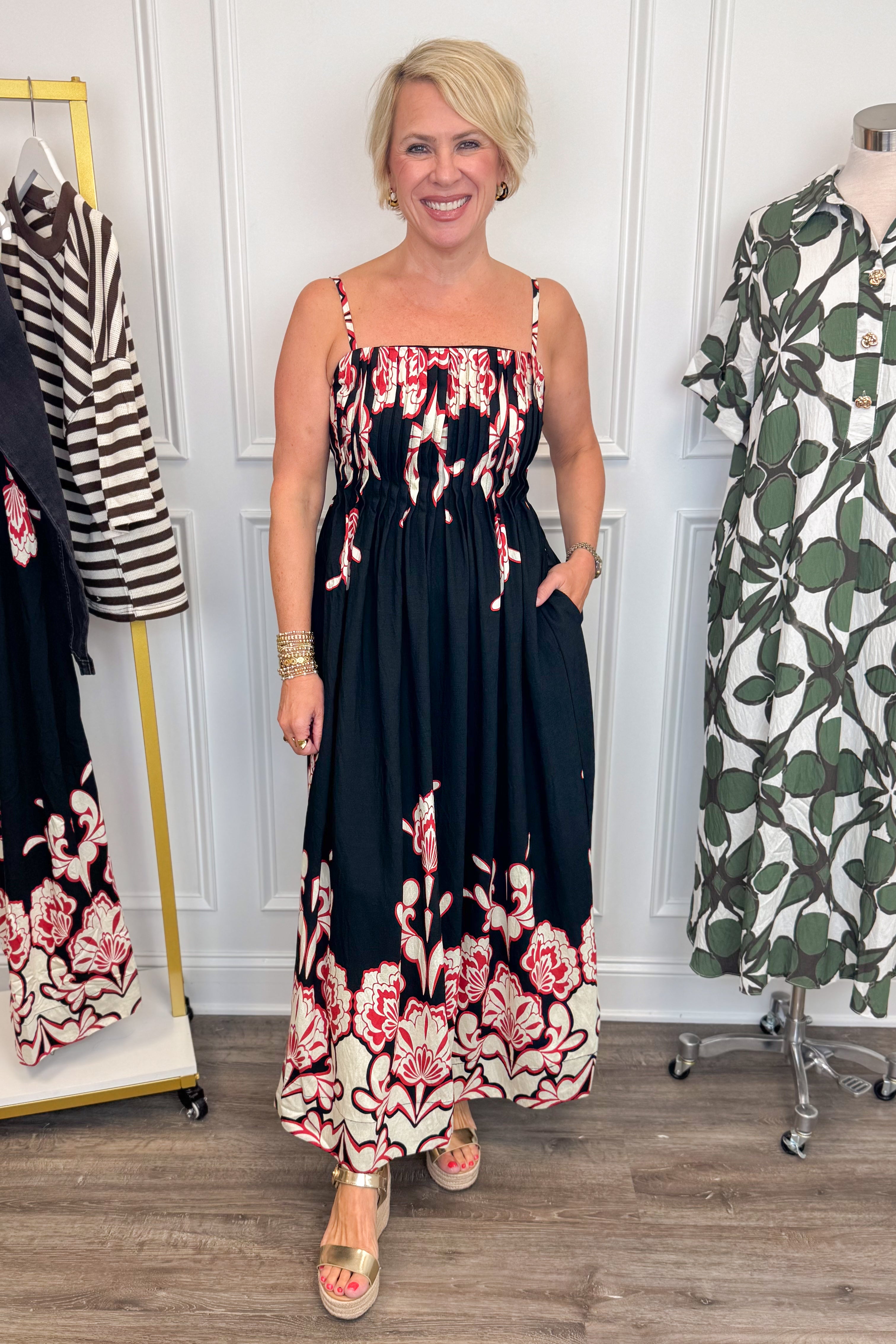 Front view of woman wearing Swagger Boutiqueās Black & Red Floral Midi Dress by Entro with sandals with clothing hanging in the background.