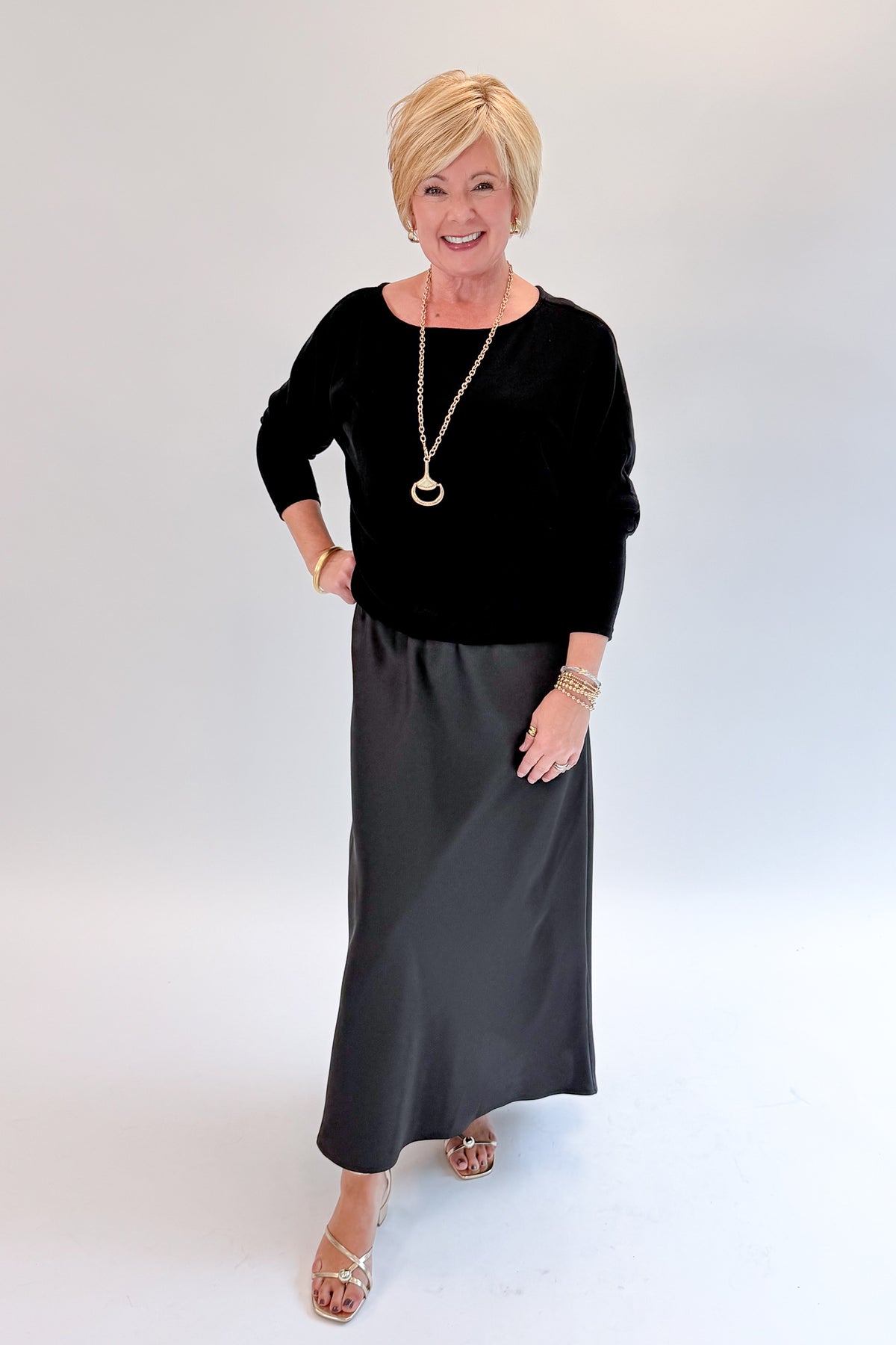 Front view of woman wearing Swagger Boutique’s Black Satin Dress and Black Layering Sweater with heels against white studio background.