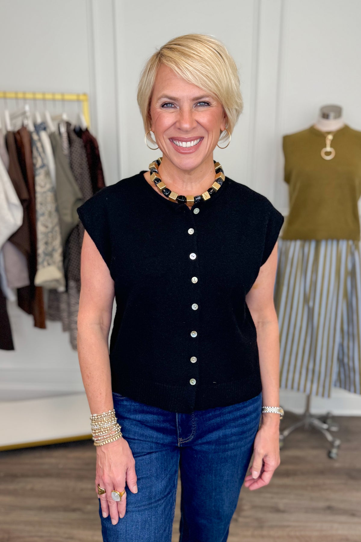 Front view of woman wearing Swagger Boutique’s Black Sleeveless Button Up Sweater with jeans with clothing hanging in the background.