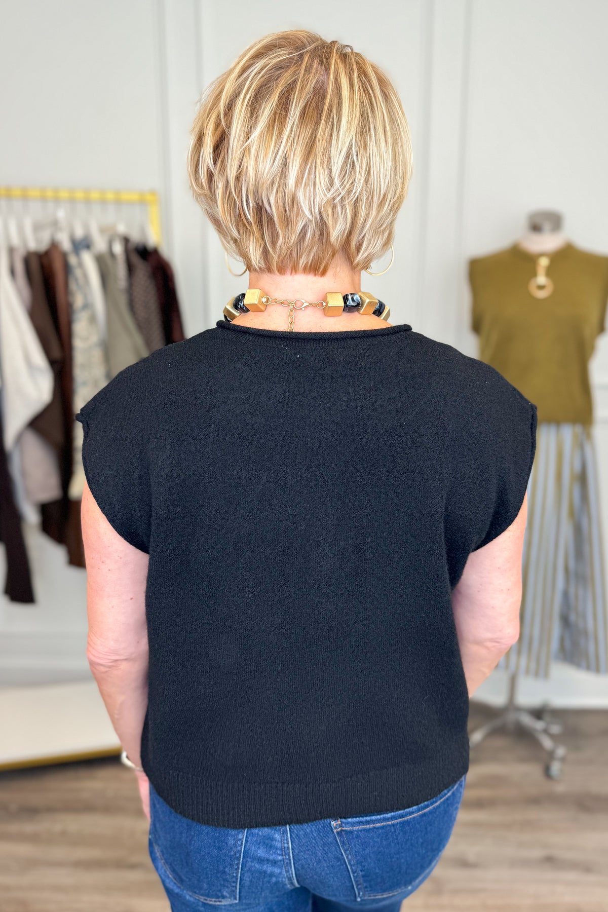 Back view of woman wearing Swagger Boutique’s Black Sleeveless Button Up Sweater with jeans with clothing hanging in the background.