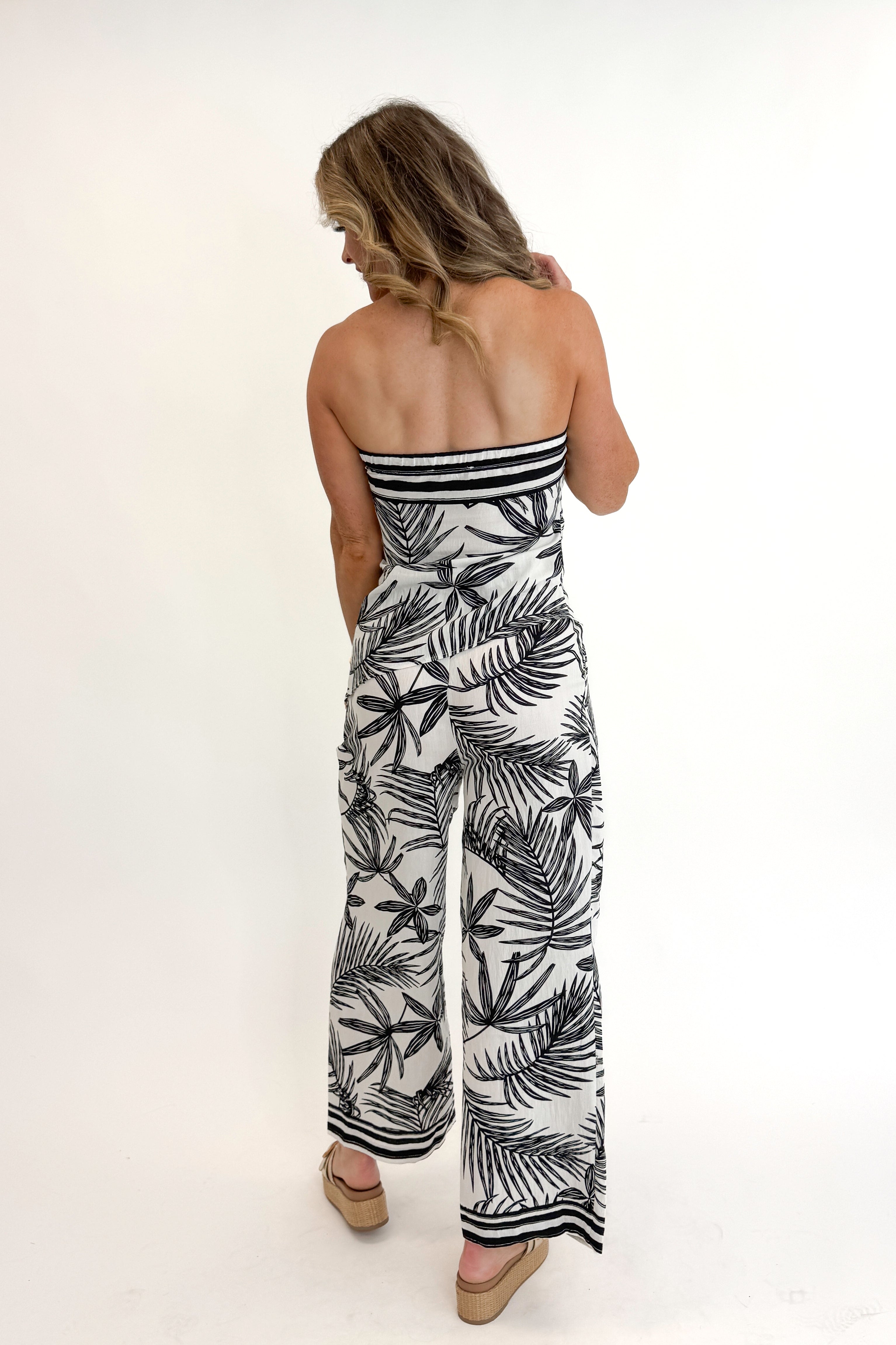 Back view of woman wearing Swagger Boutique’s Black & White Tropical Print Strapless Top with matching Black & White Tropical Print Pants against white studio background.