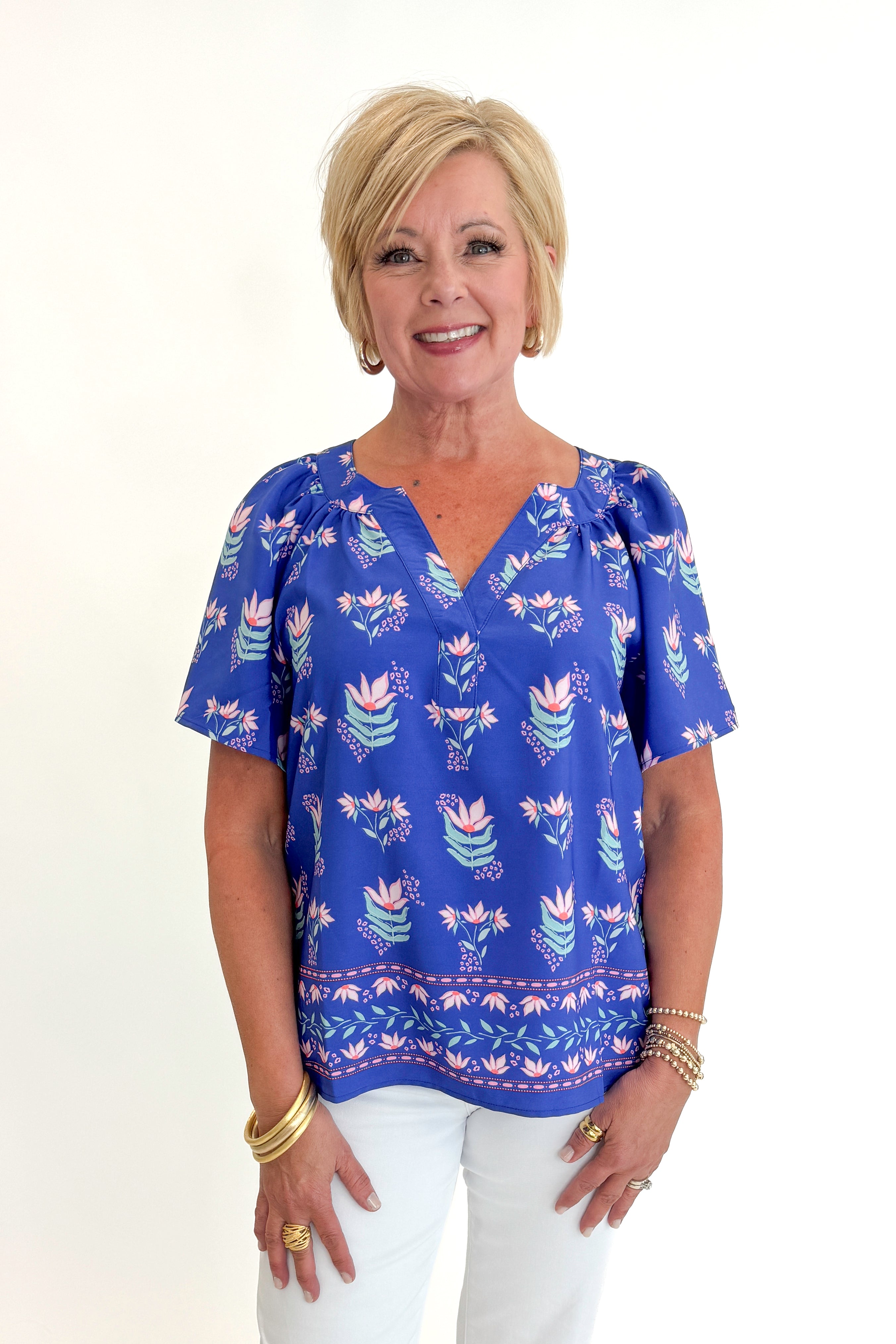 Front view of woman wearing Swagger Boutique’s Blue Petal Print Lori Top by Mary Square with white jeans against white studio background.