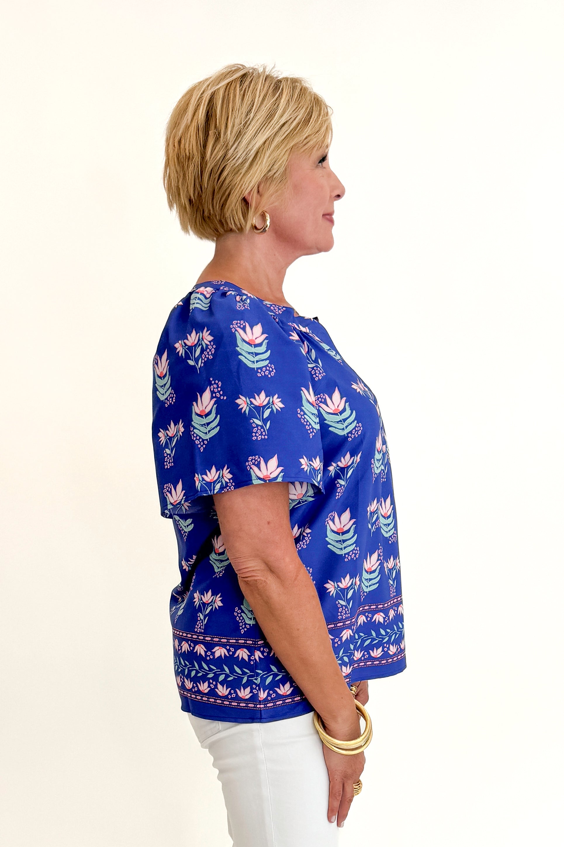 Side view of woman wearing Swagger Boutique’s Blue Petal Print Lori Top by Mary Square with white jeans against white studio background.