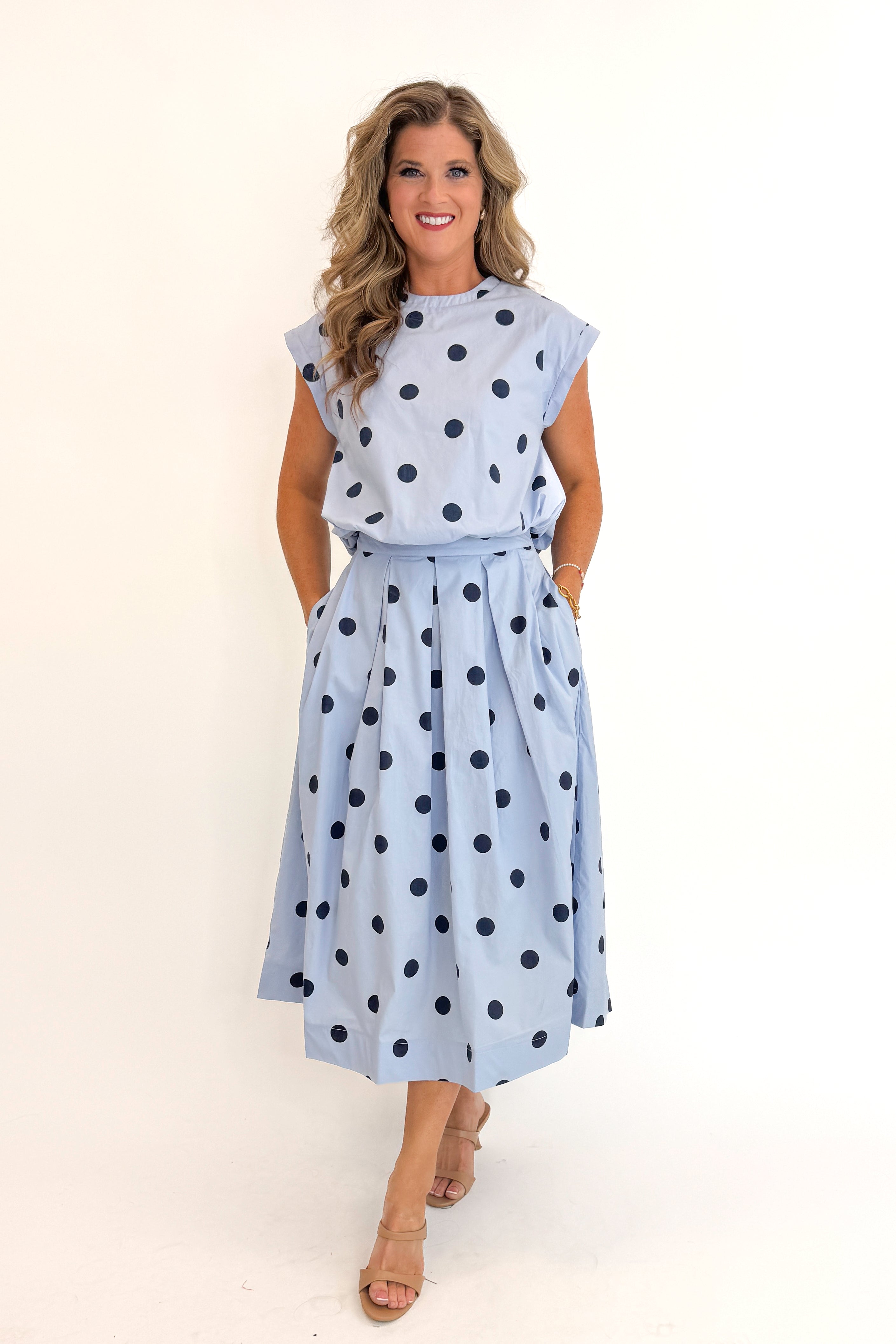 Front view of woman wearing Swagger Boutique’s Blue Polka Dot Bubble Hem Tank with matching Blue Polka Dot Pleated Midi Skirt against white studio background.