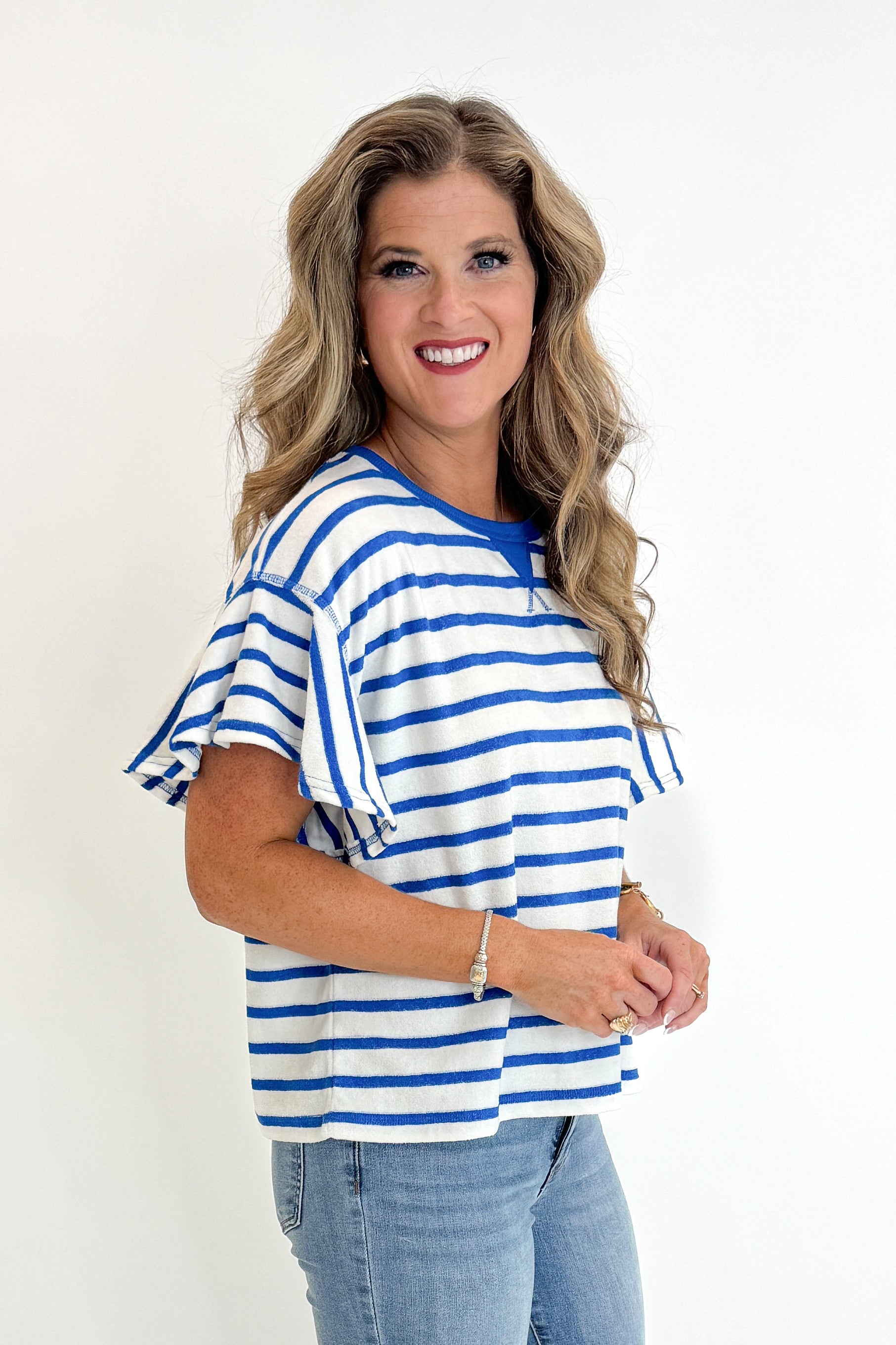 Side view of woman wearing Swagger Boutique’s Striped Flutter Sleeve Terry Top with jeans against white studio background.