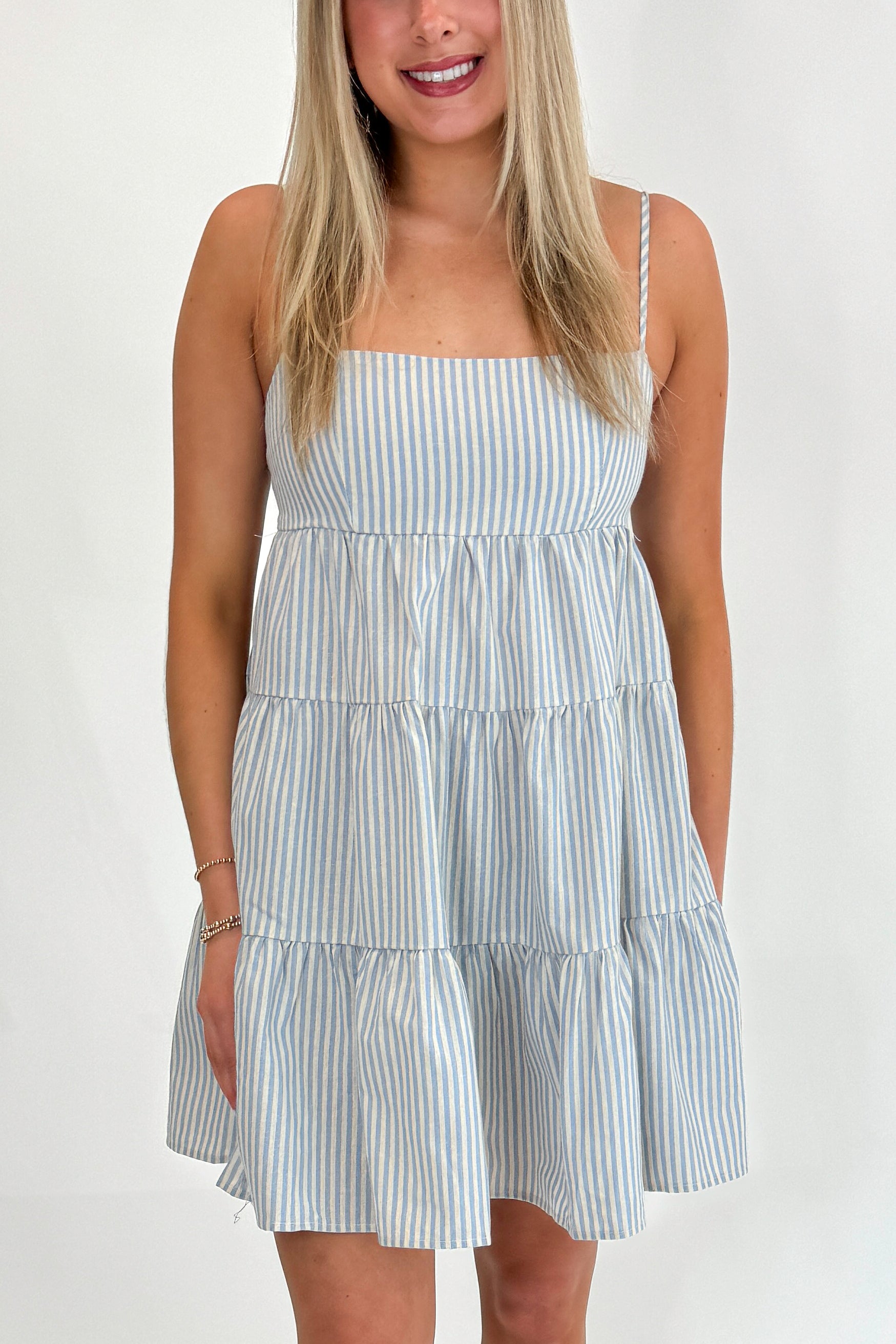 Front detail view of woman wearing Swagger Boutiqueβs Blue Striped Sleeveless Tiered Mini Dress with sandals against white studio background.