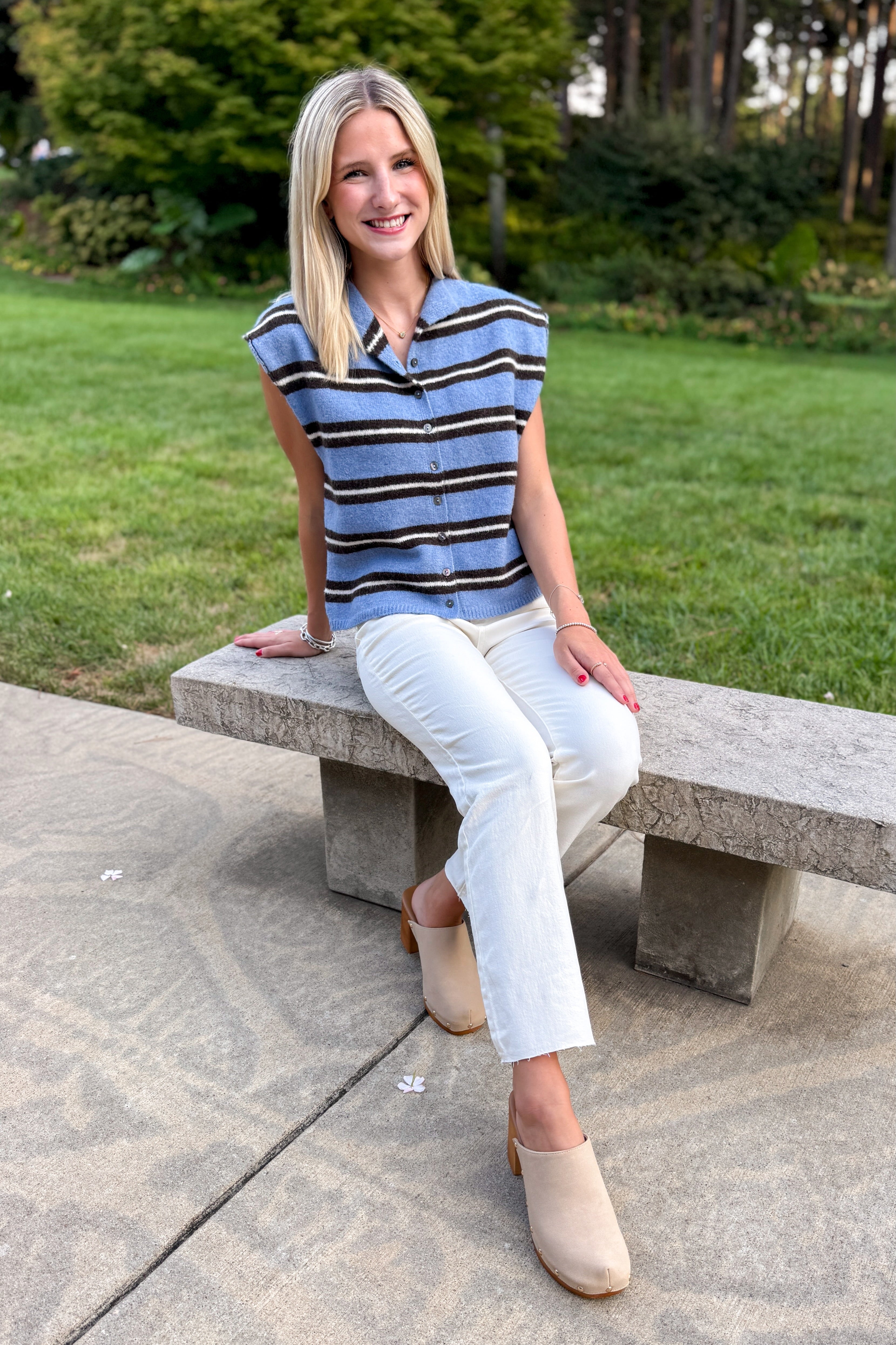 Front view of woman wearing Swagger Boutique’s Blue & Brown Striped Sleeveless Sweater by Entro with jeans sitting on a bench outside.