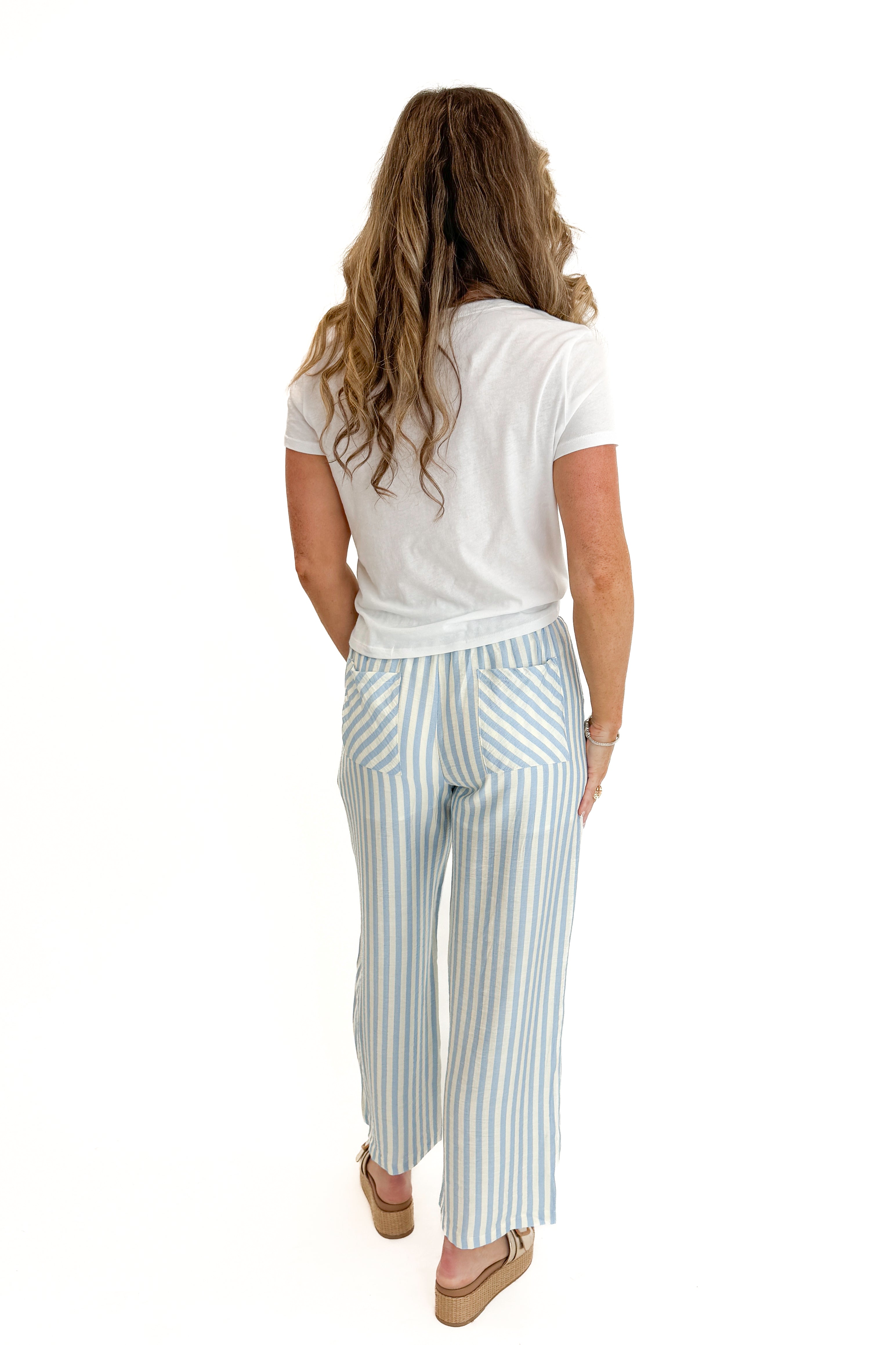Back view of woman wearing Swagger Boutique’s Blue & Cream Striped Pants with white top against white studio background.