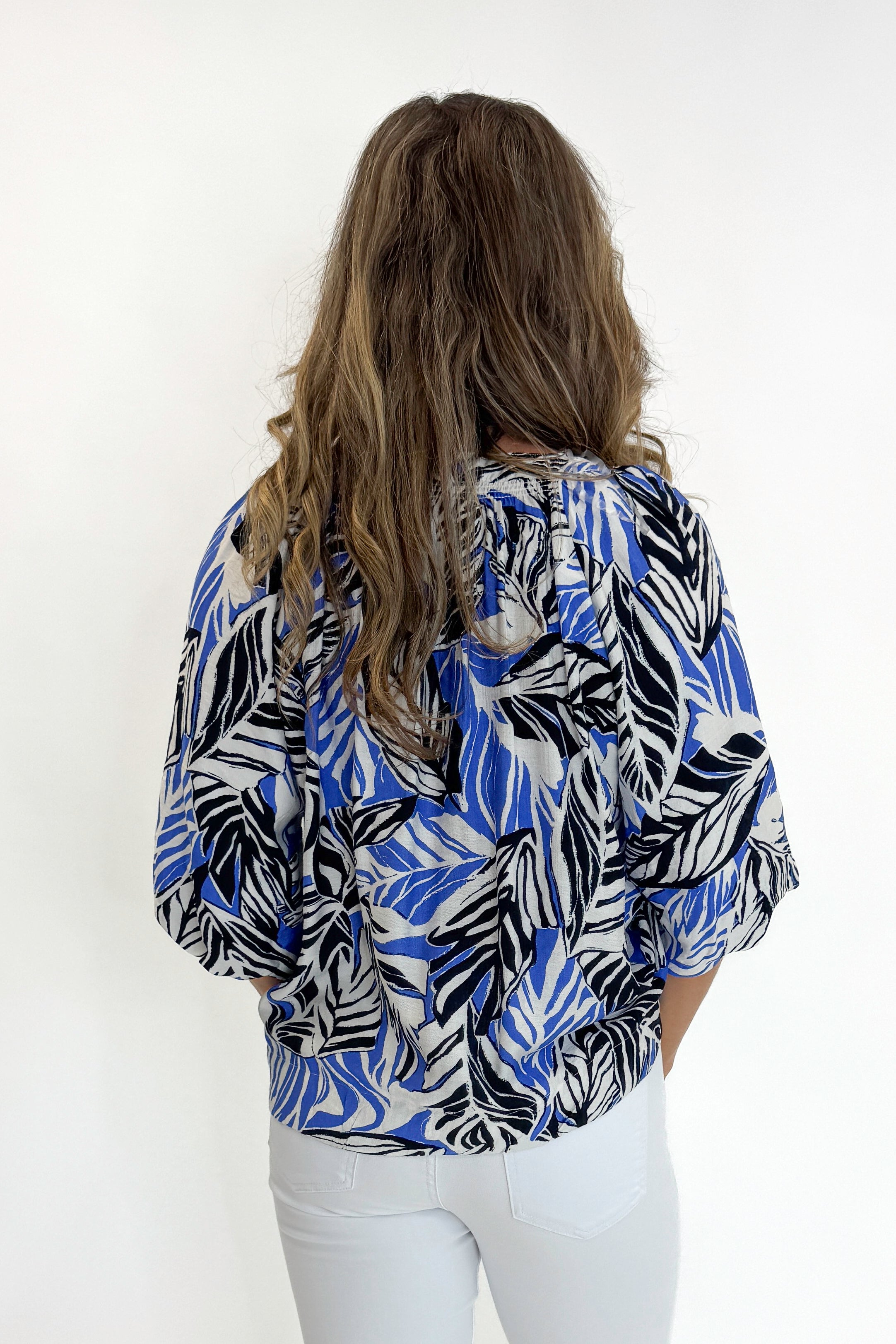 Back view of woman wearing Swagger Boutiqueās Blue & Navy Tropical Printed 3/4 Sleeve Top with jeans against white studio background.