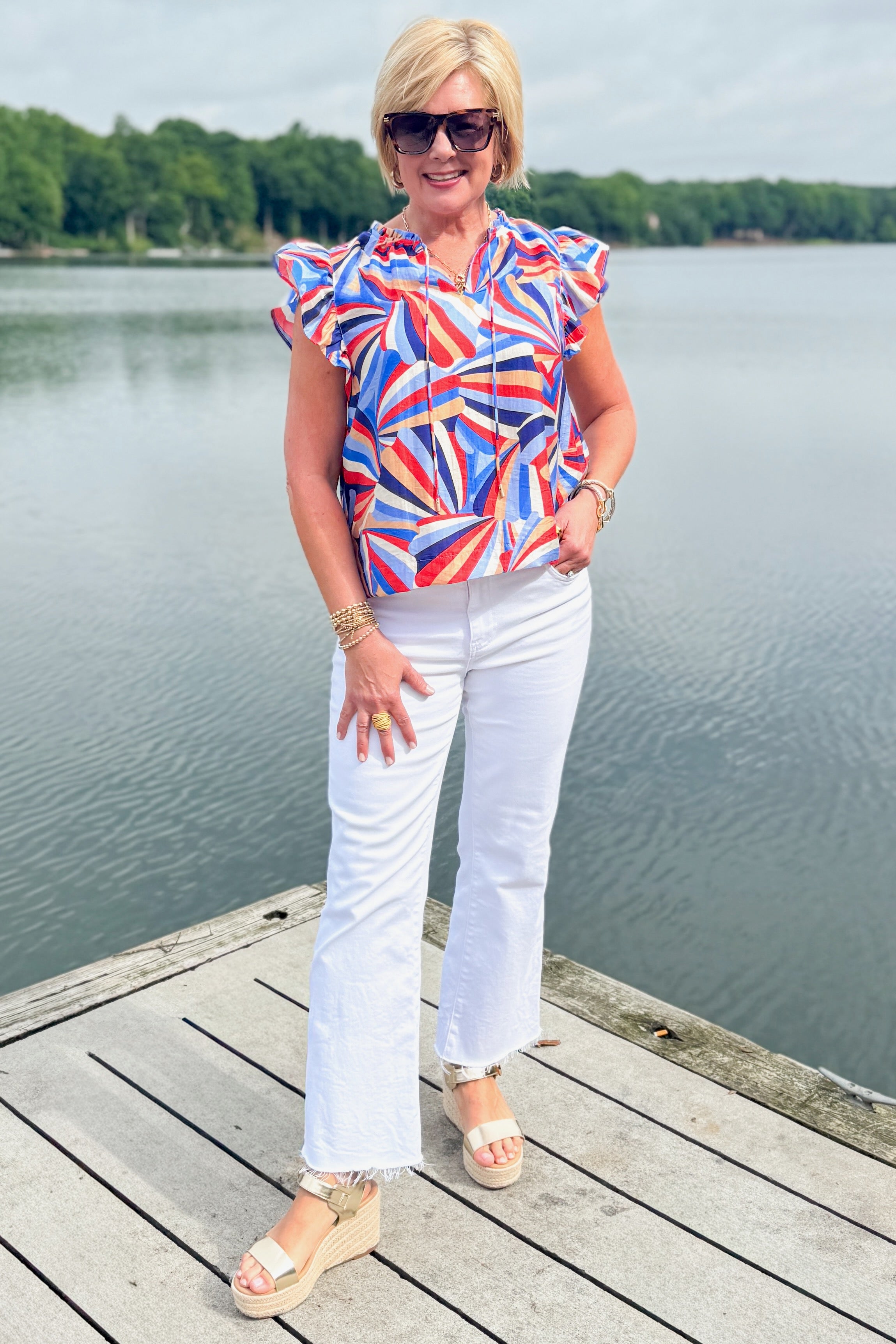 Front view of woman wearing Swagger Boutiqueās Blue & Red Printed Ruffle Sleeve Top by Skies Are Blue and jeans outside at the lake.