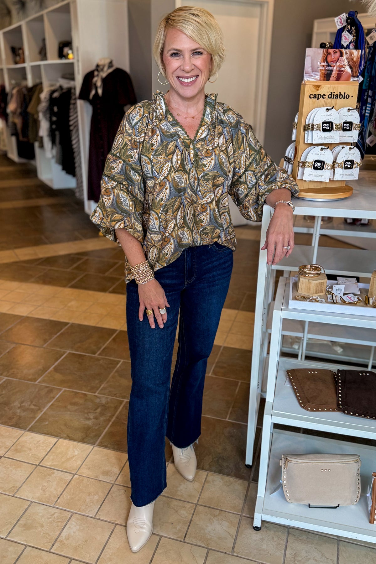 A woman stands in a boutique wearing a patterned button-up blouse with blue jeans and beige ankle boots. She is smiling and posing next to a white display shelf