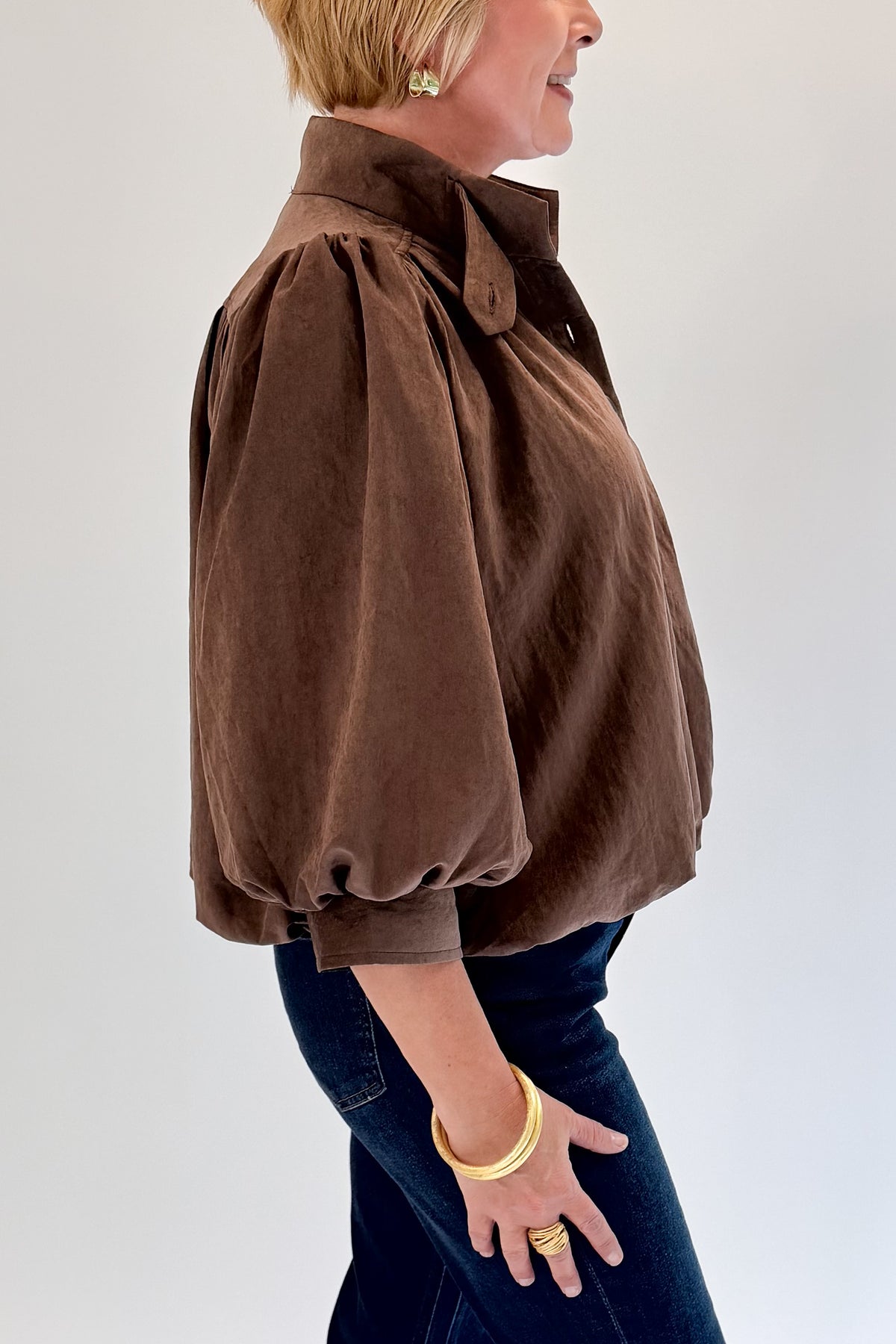 Side detail view of woman wearing Swagger Boutique’s Brown Bubble Hem Top With Buttons by Entro with jeans against white studio background.