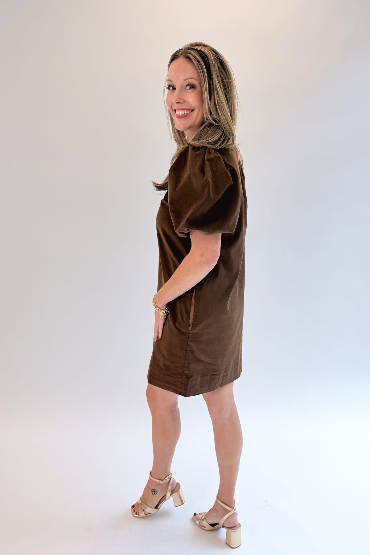 Side view of woman wearing Swagger Boutique’s Brown Embroidered Corduroy Dress with heels against white studio background.