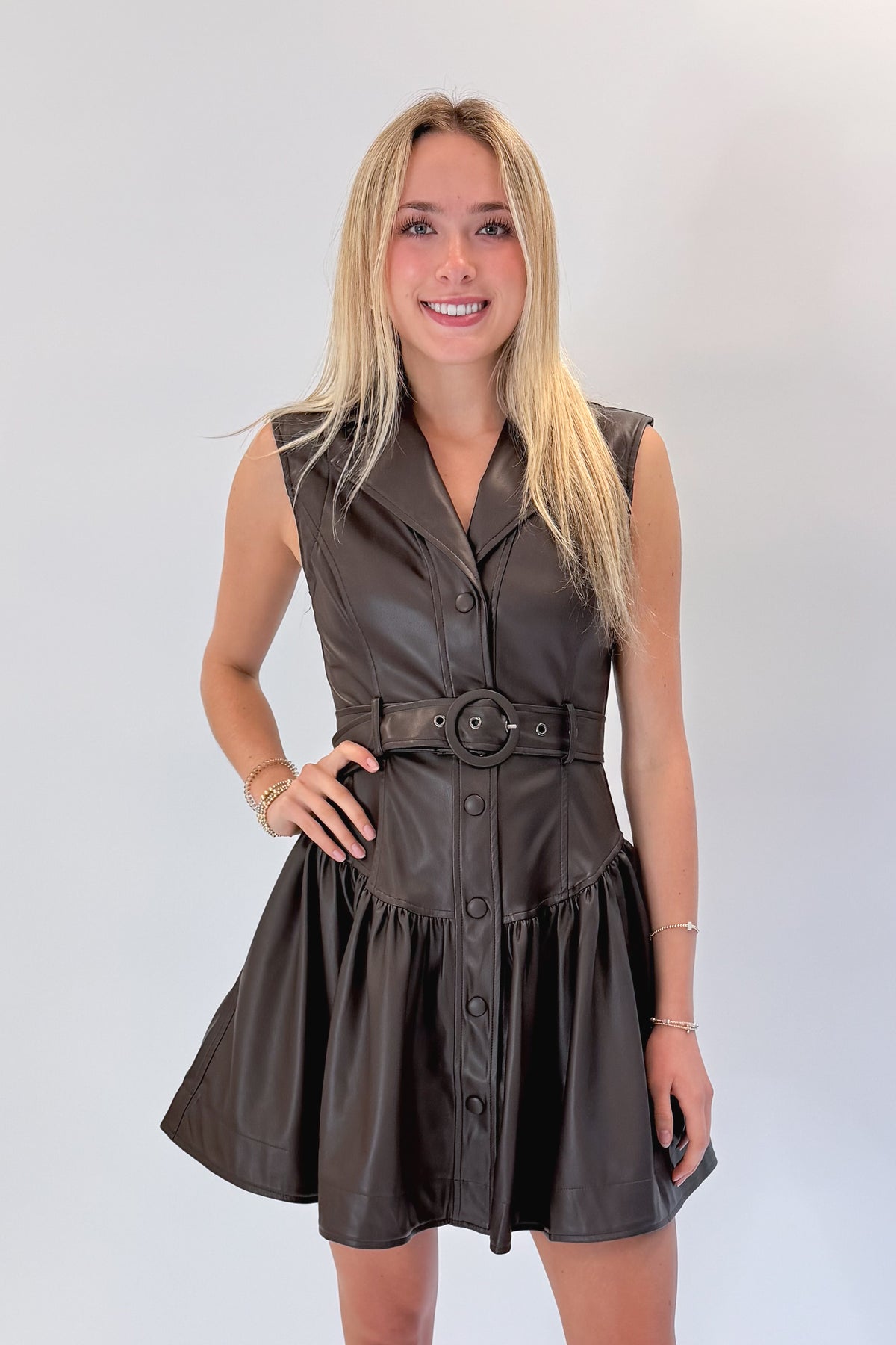 Front view of woman wearing Swagger Boutique’s Brown Faux Leather Mini Dress with heels against white studio background.