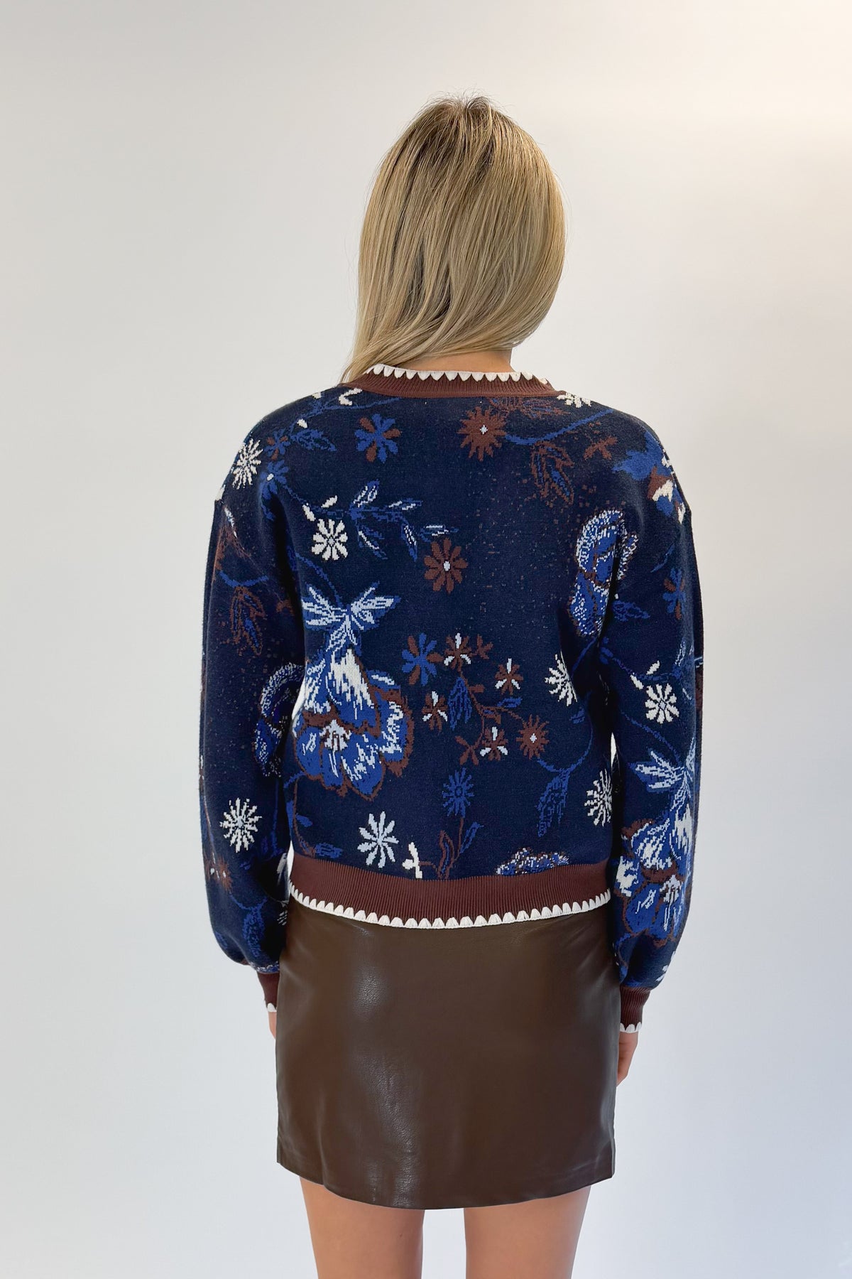 Back view of woman wearing Swagger Boutique’s Navy & Brown Printed Cardigan with Brown Faux Leather Mini Skirt against white studio background.